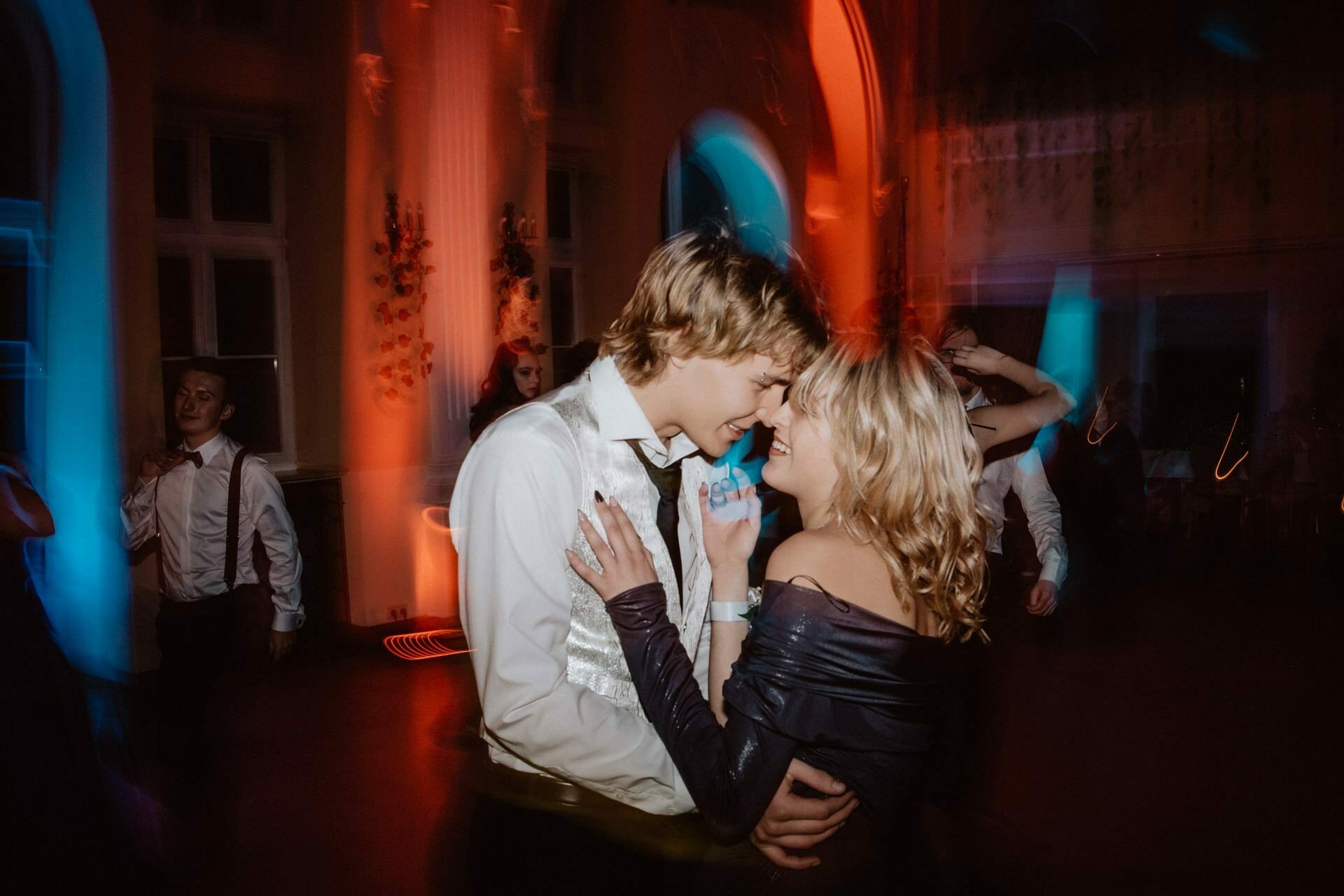 A bride and groom dance closely in dim, colorful lighting at a formal event. The man is dressed in a white shirt with a vest, and the woman in a gown with open shoulders. The other participants are visible in the background, adding to the lively atmosphere of the prom reportage.  