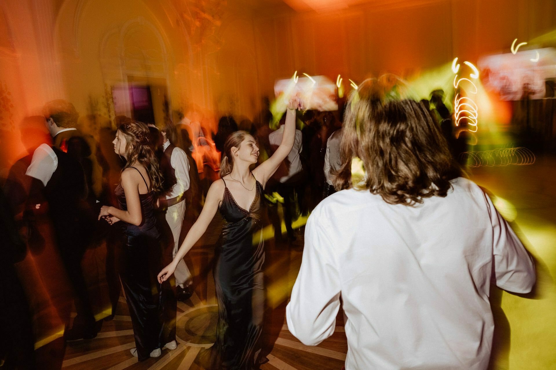 A group of people dancing at a formal event. The room is dimly lit with colored lights that create a lively atmosphere reminiscent of a prom reportage. One woman in a black dress dances with her hand raised joyfully. In the background, other participants can be seen in motion, which adds energy to the scene.   