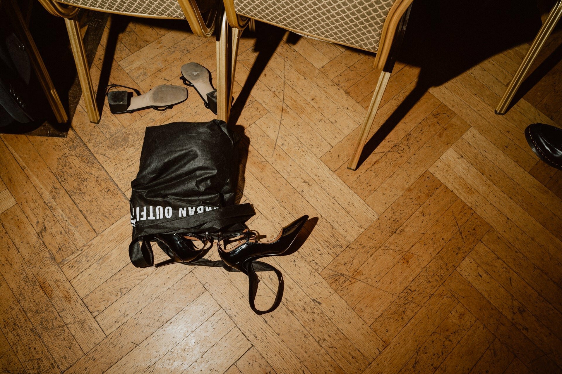 Urban Outfitters' black bag lies on the wooden dance floor next to a pair of black high-heeled shoes, beige sandals and the legs of several chairs with light brown cushions - a scene straight out of a prom report.