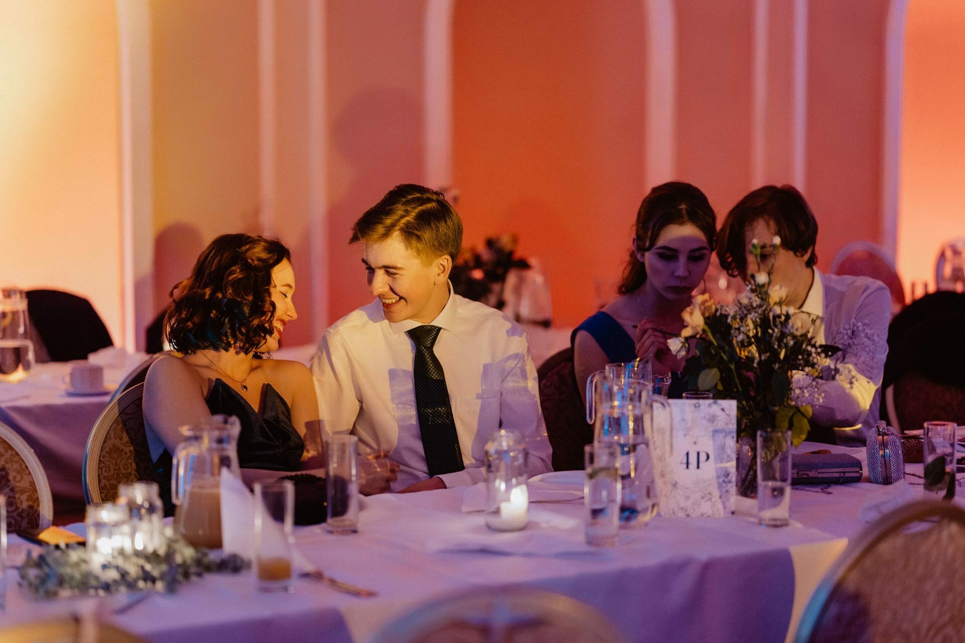 During a reportage of a prom, four young people dressed in semi-formal attire sit at a decorated table. The two on the left are smiling at each other, while the two on the right are focused on their activities. The table is decorated with flowers, candles and the table number "4.  