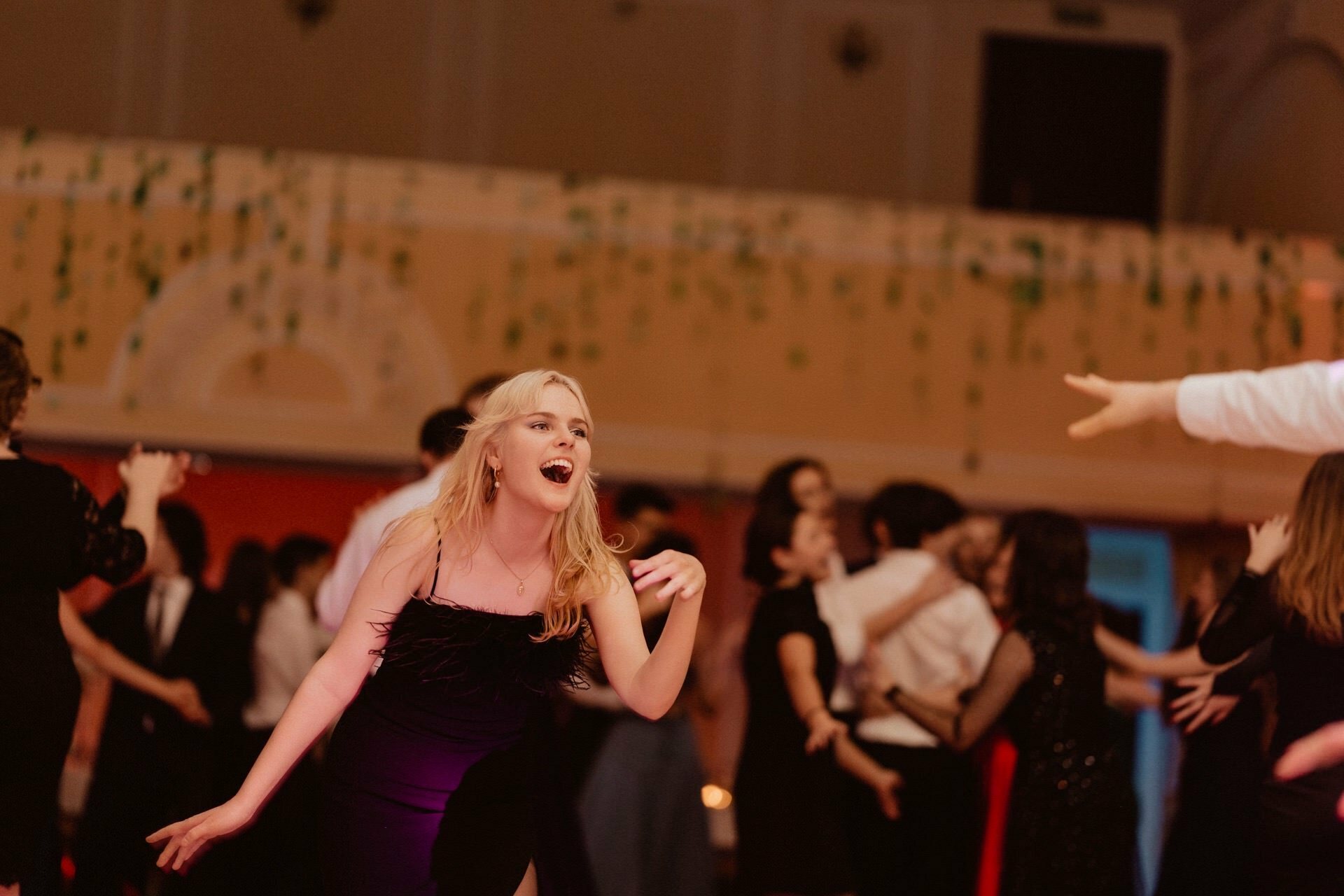 A blonde woman in a black dress dances and sings joyfully at a lively party in a hall that resembles a bustling report from a prom. Around her, other people are also dancing and chatting. The background is decorated with hanging green strings, and the atmosphere seems festive and warm.  