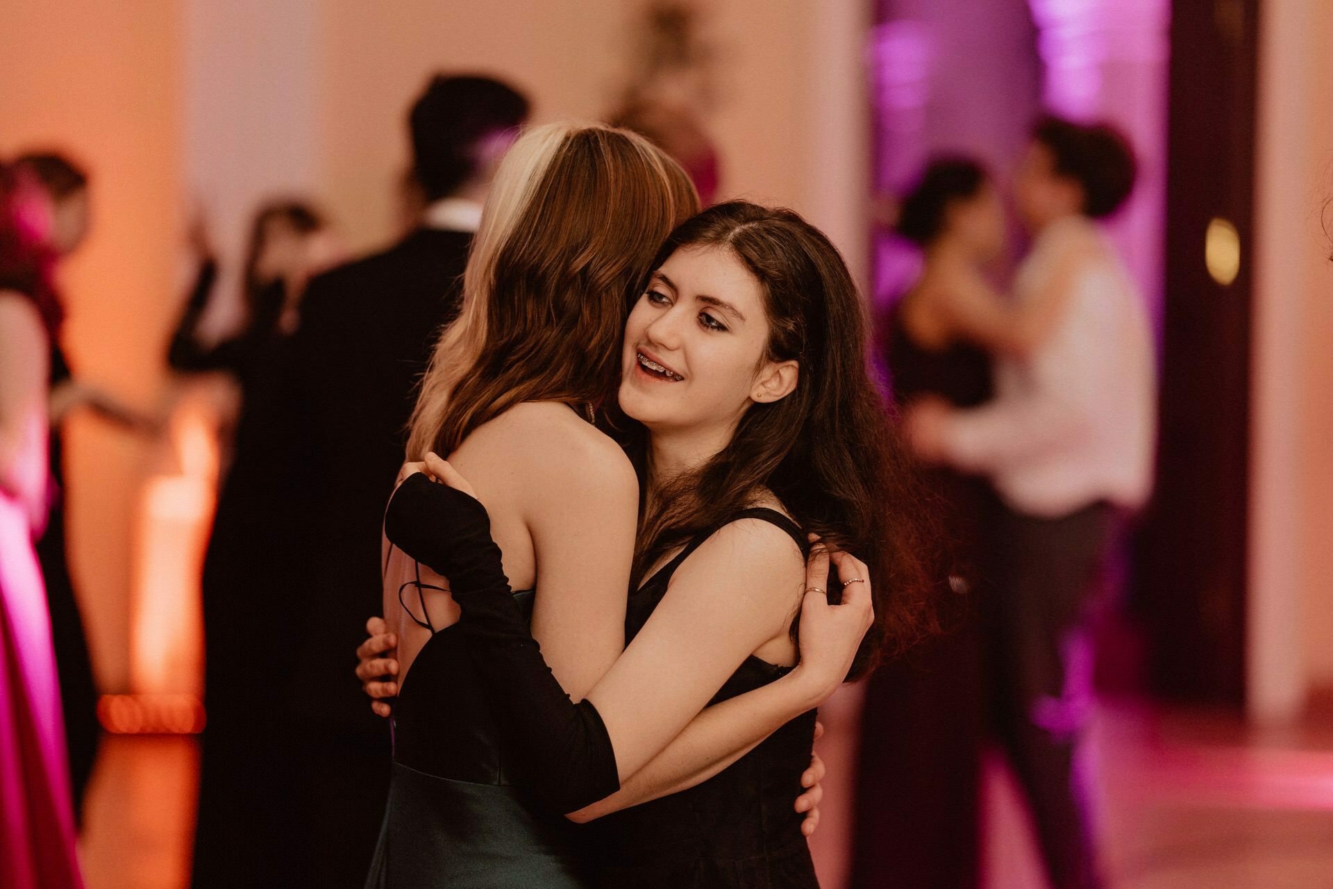 Two women embrace each other warmly as they dance in a softly lit room filled with other couples. The background is softly blurred, highlighting the colorful lights and lively atmosphere, reminiscent of a vibrant prom reportage. 