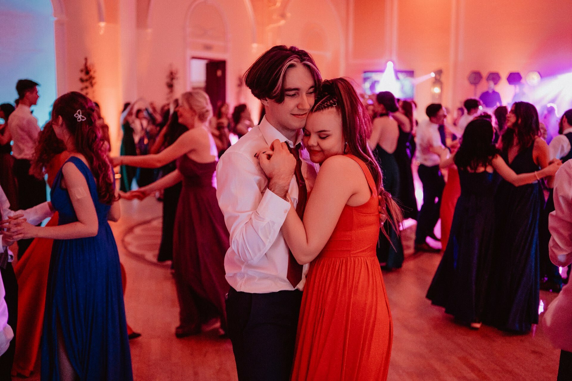 The newlyweds embrace and smile as they dance close in the warmly lit ballroom filled with people. The girl is dressed in an airy red dress and the boy in a white shirt with a patterned tie. The other guests of the prom reportage are also dancing and enjoying the festive atmosphere.  