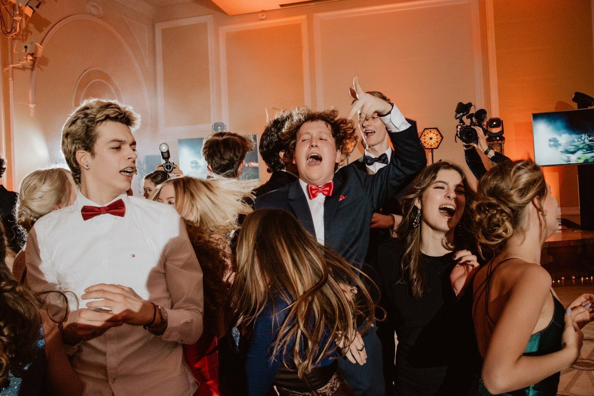 A group of young people enthusiastically dancing at a formal party, perhaps as part of a reportage of a prom. They are dressed in suits and dresses, some wearing bow ties. The crowd seems lively and joyful, with several people smiling, laughing and raising their hands in excitement. Photographers in the background capture the moment.   