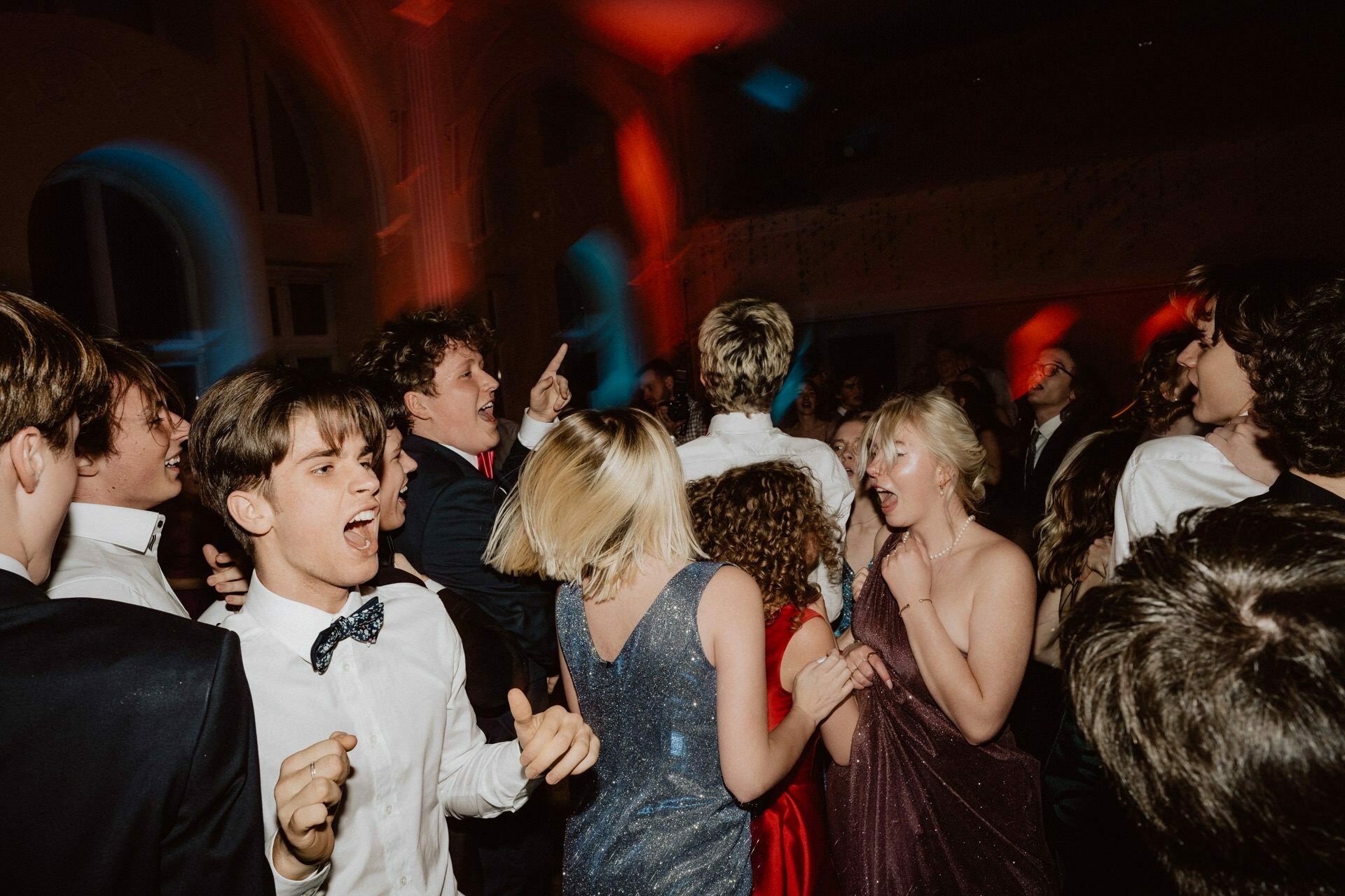 A lively group of young adults dance vigorously during what resembles a prom. The room is dimly lit with colored lights, creating a festive atmosphere. Everyone seems to be enjoying the moment, smiling and interacting with each other. Among the participants, formal attire is the most important.   