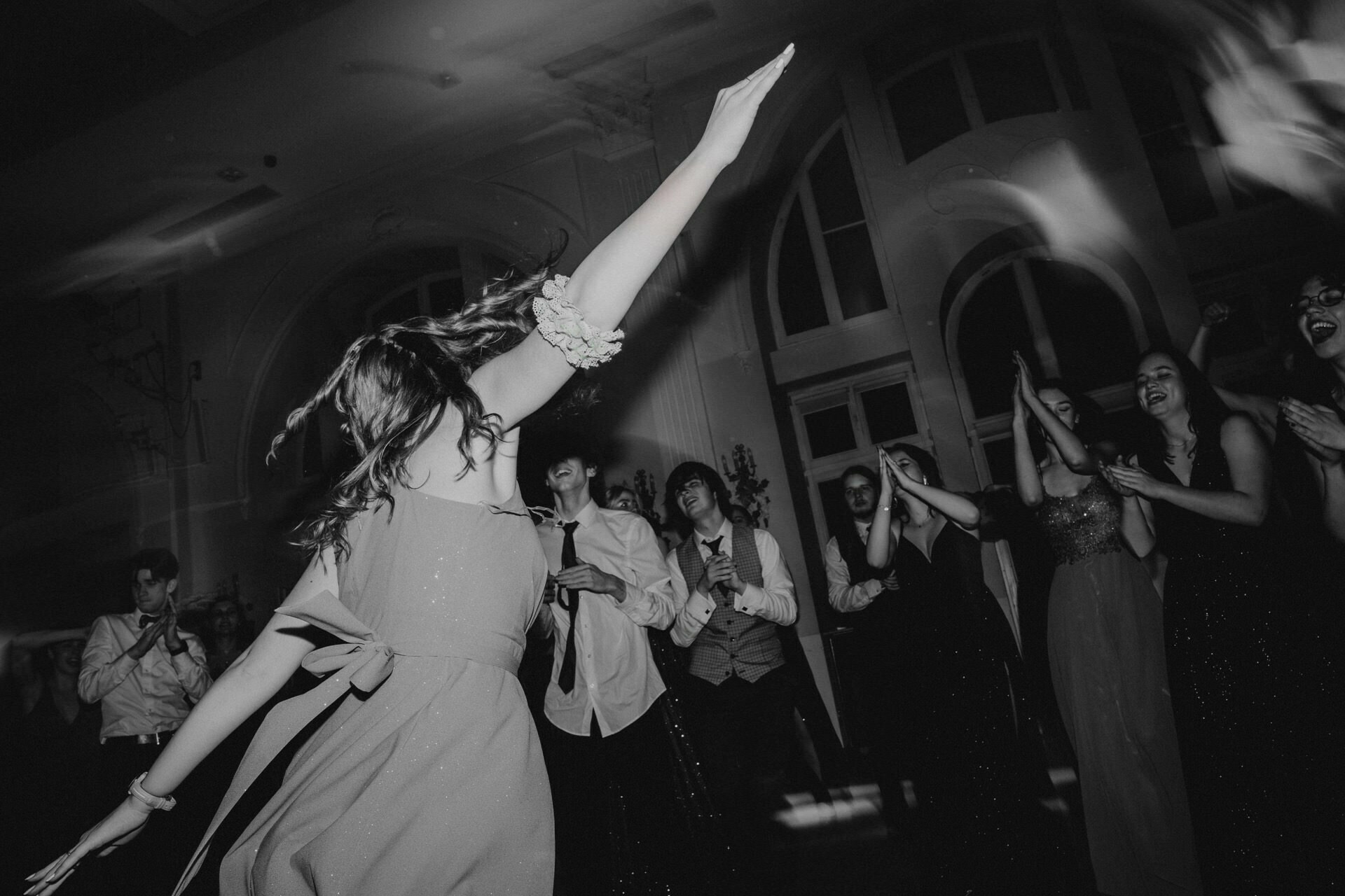 The black-and-white photo shows a vibrant scene in "Report from the Prom." A person with long hair in the foreground is dancing with his hands raised. In the background, other people dressed in formal attire are clapping, cheering and looking at the dancer with smiles.  