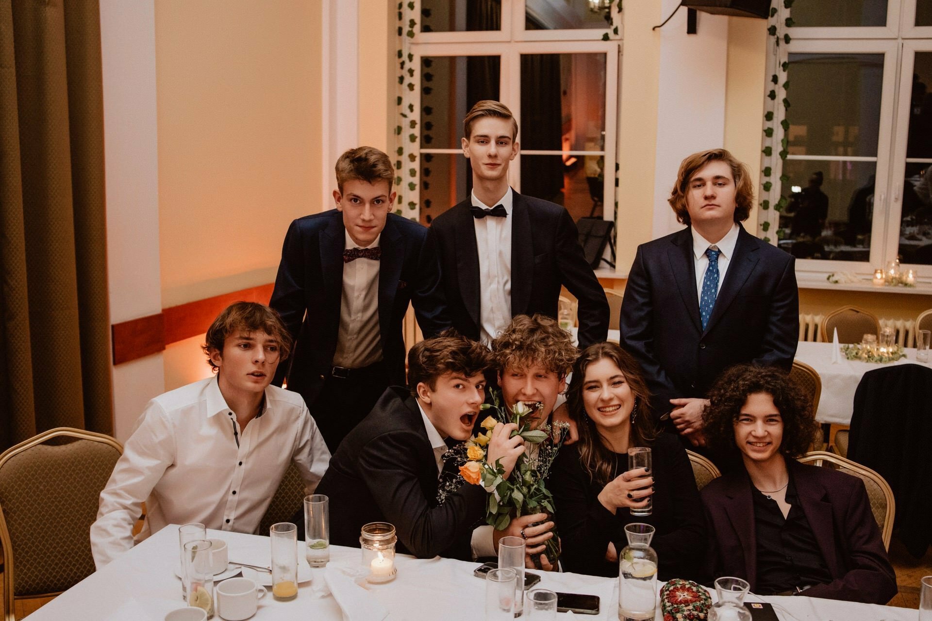 A group of eight young people posing together at a party in a room. They are dressed in formal attire, some sitting, others standing behind them. Several are holding drinks, and one person is nibbling on a bouquet of flowers for fun. The setting includes a large window and decorated tables that capture the atmosphere of a prom reportage.   