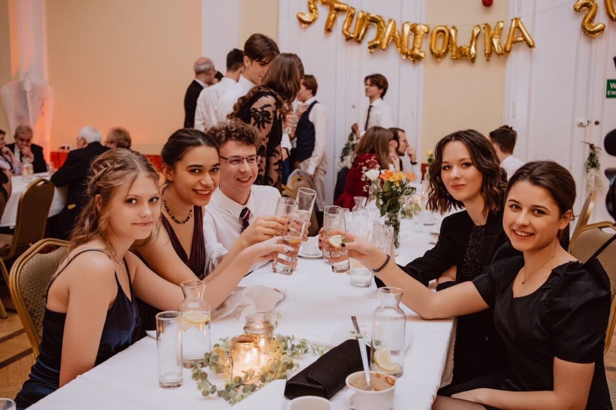 Grupa młodych ludzi ubranych w formalne stroje siedzi wokół stołu i wznosi toast drinkami w dłoniach. Stół udekorowany jest świecami i kwiatami. W tle złote balony z napisem „Studniówka”. Można zobaczyć mieszających się gości. Atmosfera uchwycona w tym reportażu ze studniówki jest świąteczna.