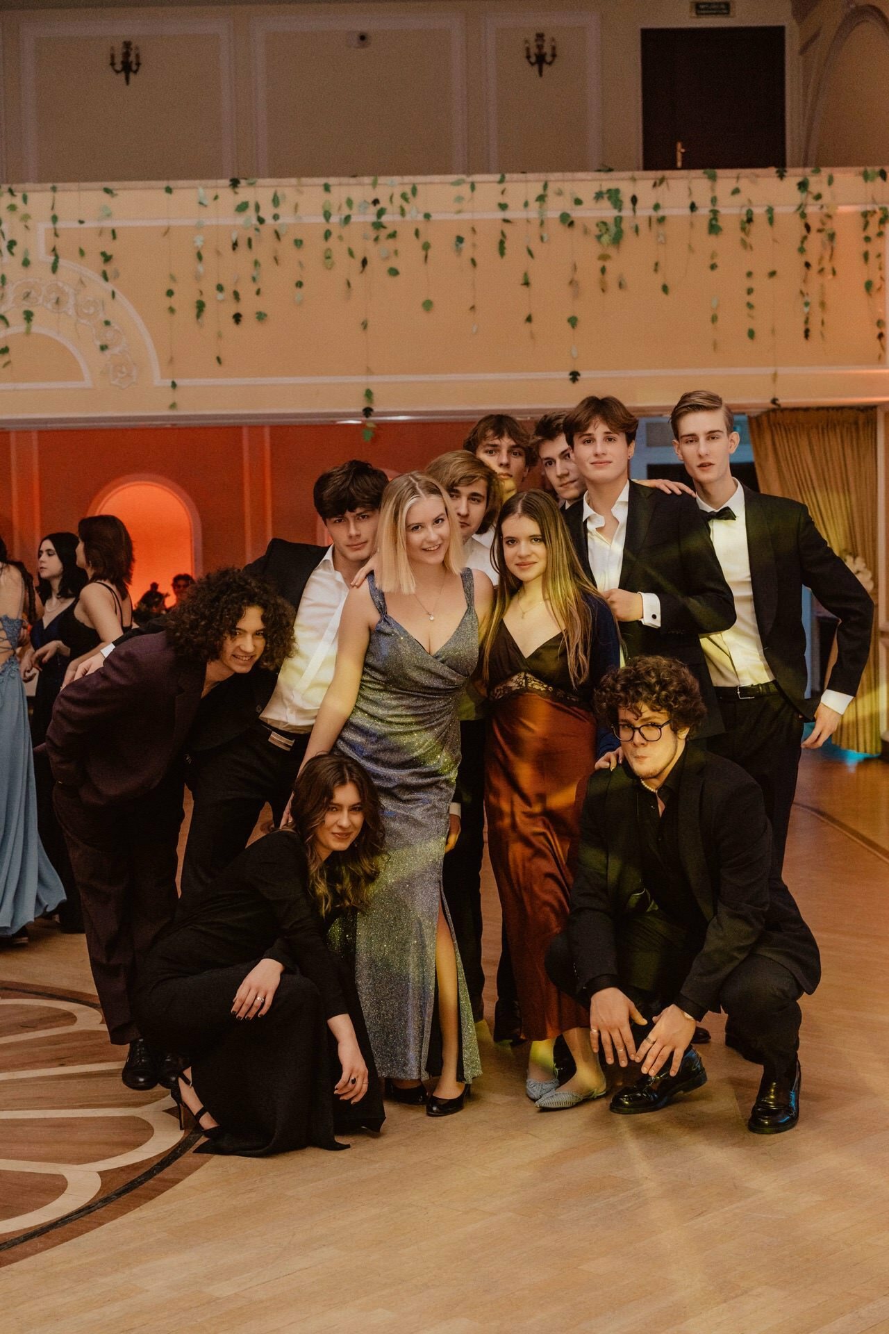 A group of young adults dressed in formal attire pose together in a ballroom for a prom reportage. Some are dressed in suits, others in gowns. They stand and kneel in a semicircle, smiling and enjoying the event. In the background, elegant decor and hanging greenery.   