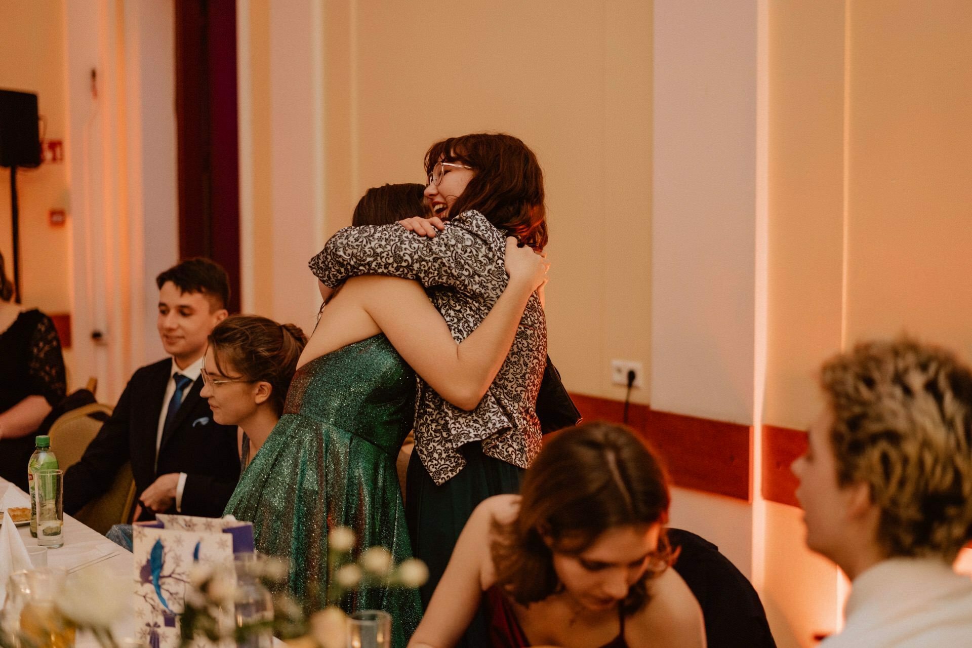 Two people embrace warmly at a festive gathering reminiscent of a prom report. Several people sit at a table decorated with flowers, immersed in conversation and laughter. The room is softly lit, creating a cozy and festive atmosphere.  