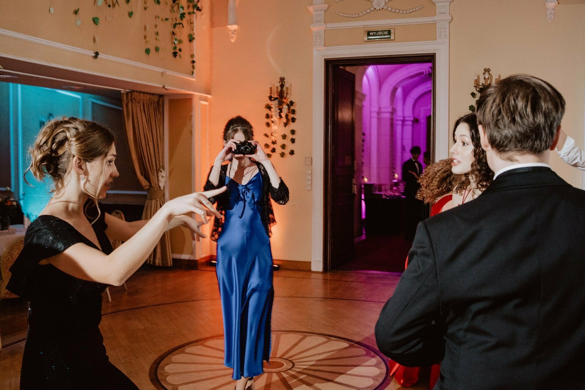 A group of people at a formal party are dancing and enjoying themselves in a well-appointed room with colorful lighting. One person in a blue dress captures the moment with a camera, giving it the appearance of a vivid *wedding prom reportage*, while others in elegant attire circle around her. 