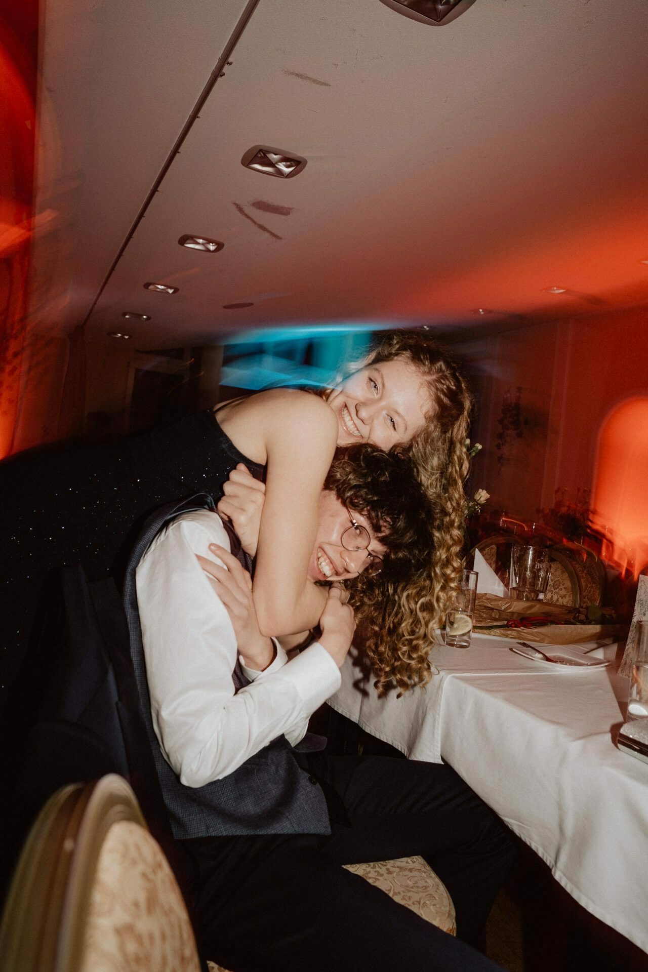 A young woman with curly blond hair playfully hugs a young man with curly brown hair from behind, both smiling and formally dressed. They sit at a table with a white tablecloth and glass, in a warmly lit space with a festive atmosphere, capturing the joyful mood of the "prom reportage." 