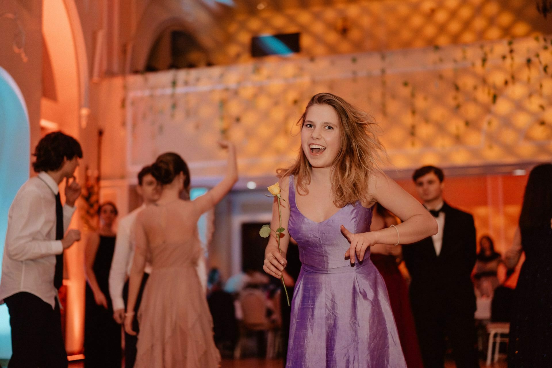 A young woman in a purple dress smiles and holds a single rose while dancing at an elegant event. The prom reportage shows others, dressed in formal attire, also dancing in a softly lit venue with intricate architecture and warm lighting in the background. 