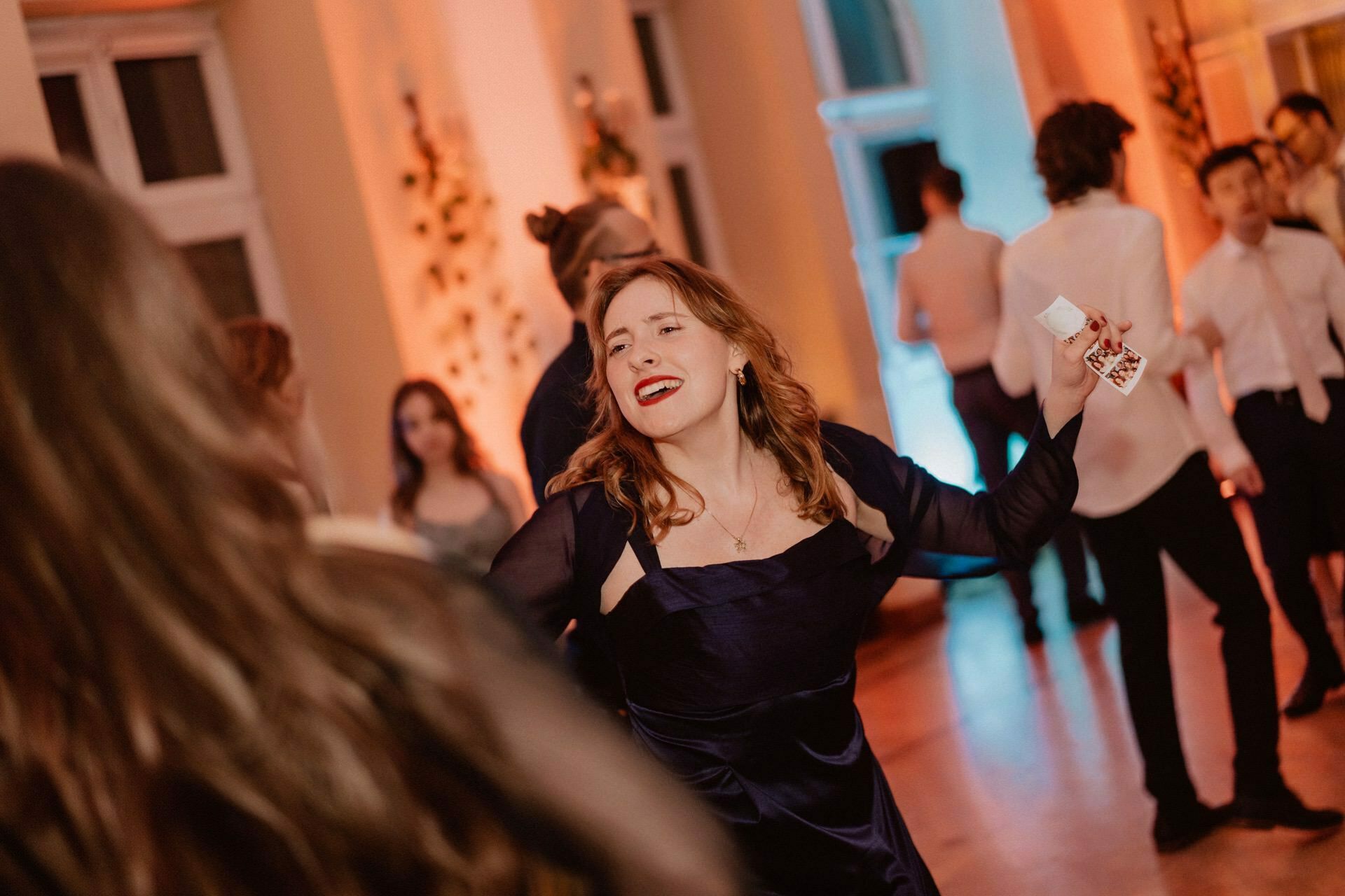 A woman in a dark blue dress smiles and dances during a lively event in the room, surrounded by other dancing guests. The room is warmly lit with a mix of orange and blue lights, adding to the festive and joyful atmosphere reminiscent of a vibrant prom reportage. 