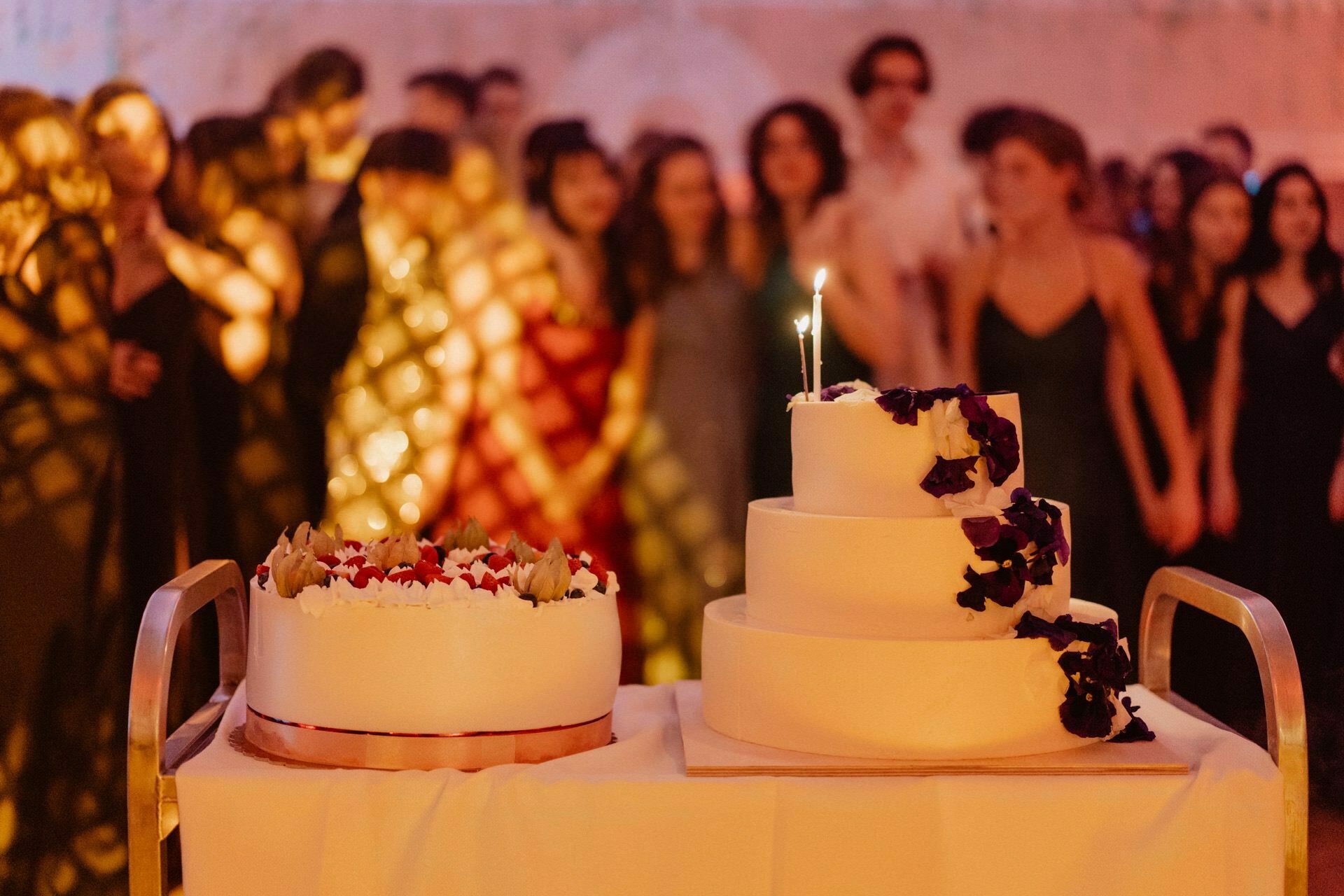 In the reportage from the prom you can see two decorated cakes; one is round with flowers on top, and the other is a three-tier cake decorated with purple and white flowers. A lit candle decorates the top level. In the background stands a group of people dressed in formal attire, somewhat fuzzy.  