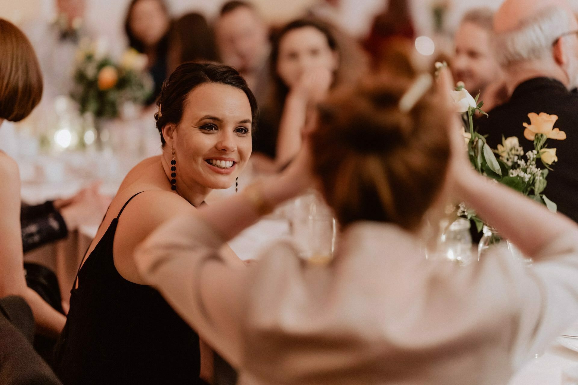 A woman in a black dress, with short hair and black earrings smiles warmly at another woman with pinned up red hair, with her hands raised in the air, creating an animated scene reminiscent of a reportage from a prom, at a candlelit wedding reception with guests blurred in the background.