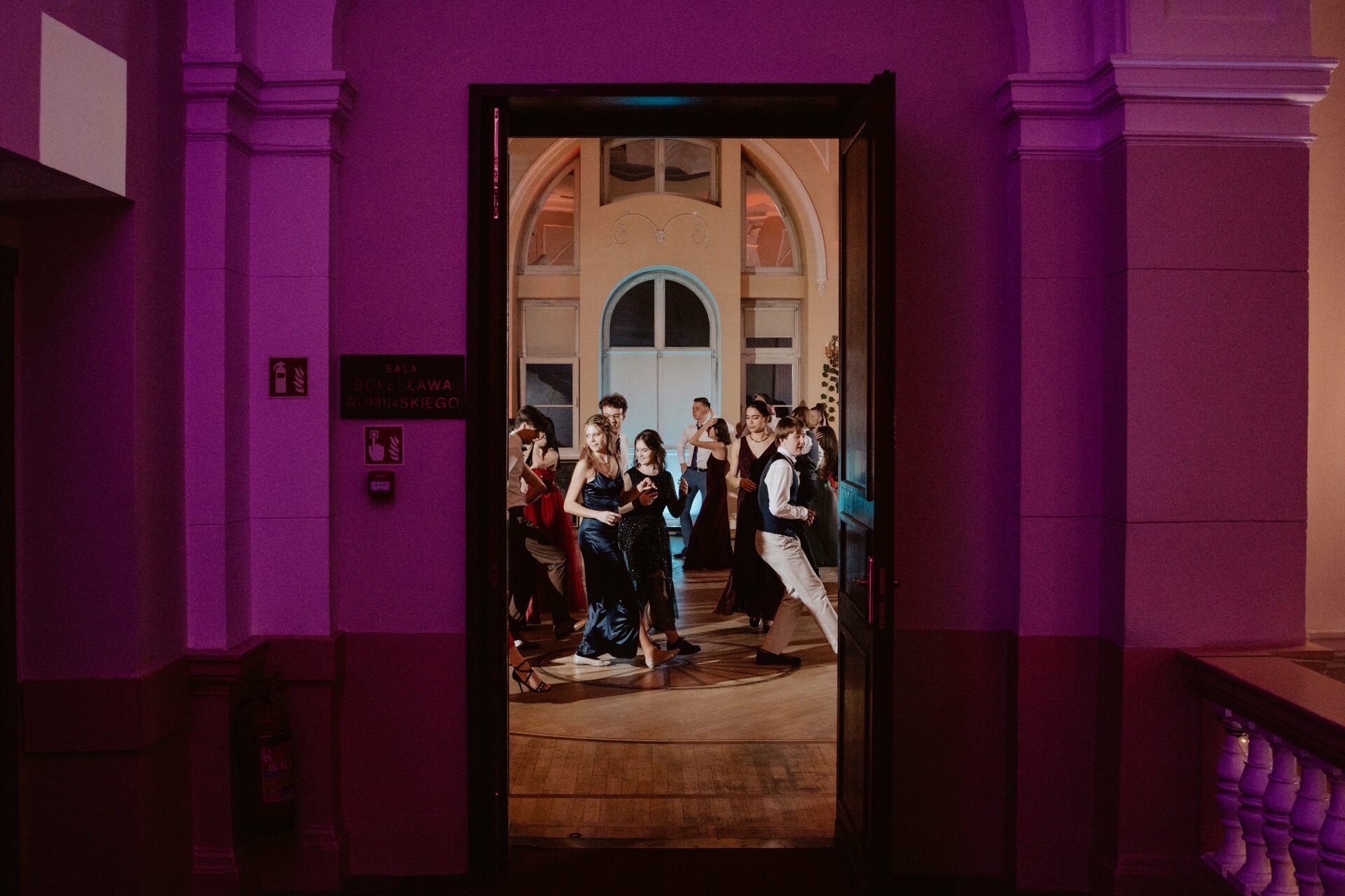 A group of formally dressed people dance enthusiastically in a warmly lit room with high ceilings and arched windows. The scene, reminiscent of a vivid reportage from a prom, is viewed through an open doorway, while the purple lighting of the hallway creates a colorful contrast. 