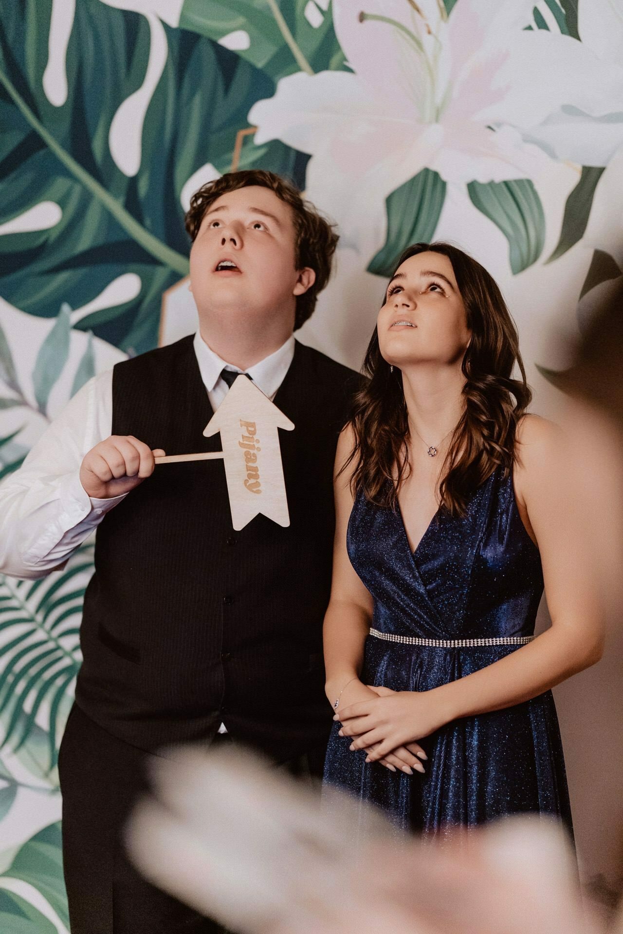 A young man in a formal vest and tie holds a small wooden arrow sign, standing next to a young woman in a blue dress. Both look up with curious faces. Behind them, referring to a prom reportage, is a decorative background with large green leaves and white flowers.  