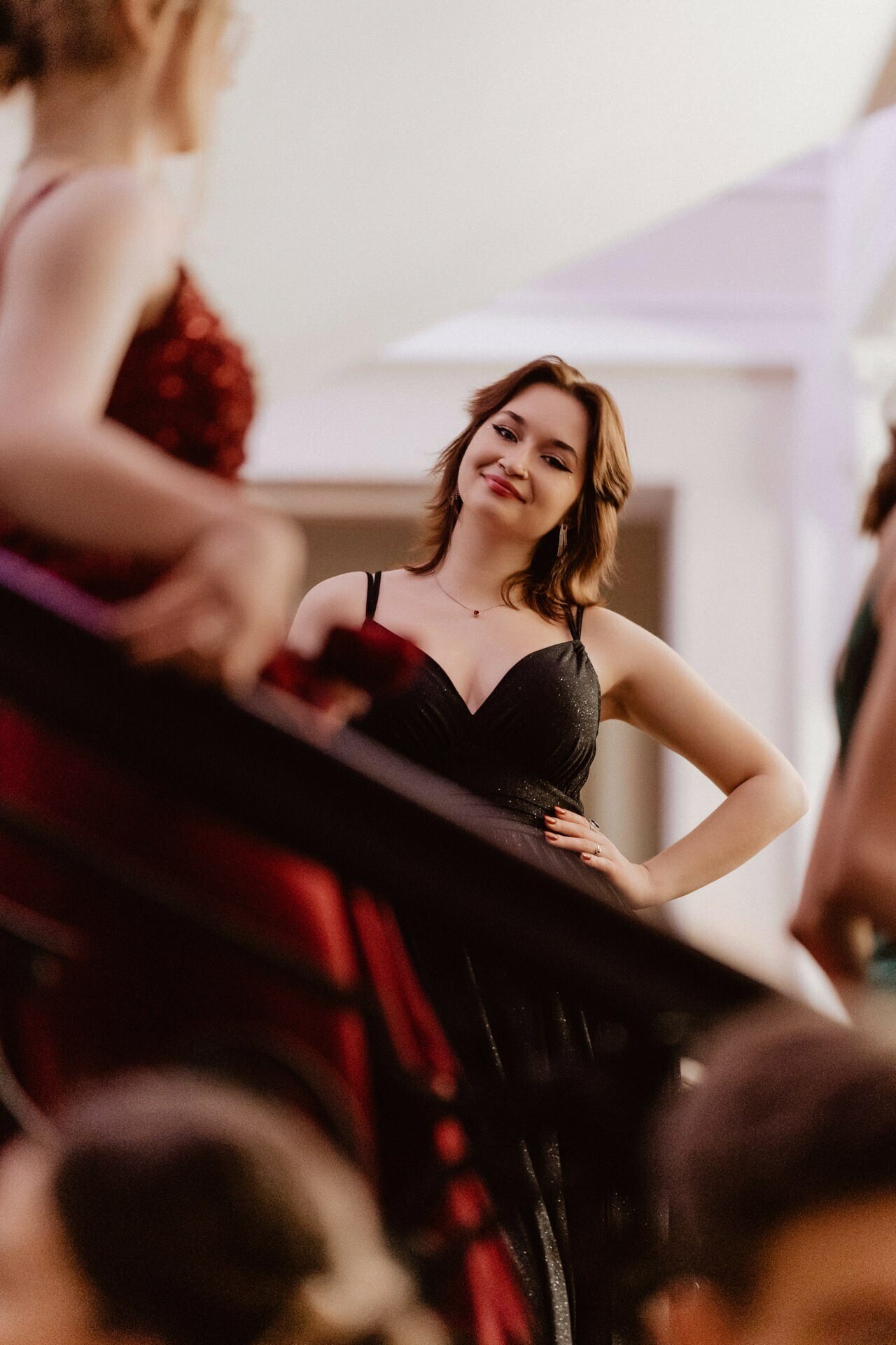 A woman dressed in a black dress stands confidently at the bottom of the stairs, smiling, holding her hand on her hip. She looks at another person in a red dress, who is partially visible in the foreground. The scene suggests an elegant setting, reminiscent of "Prom Report.  