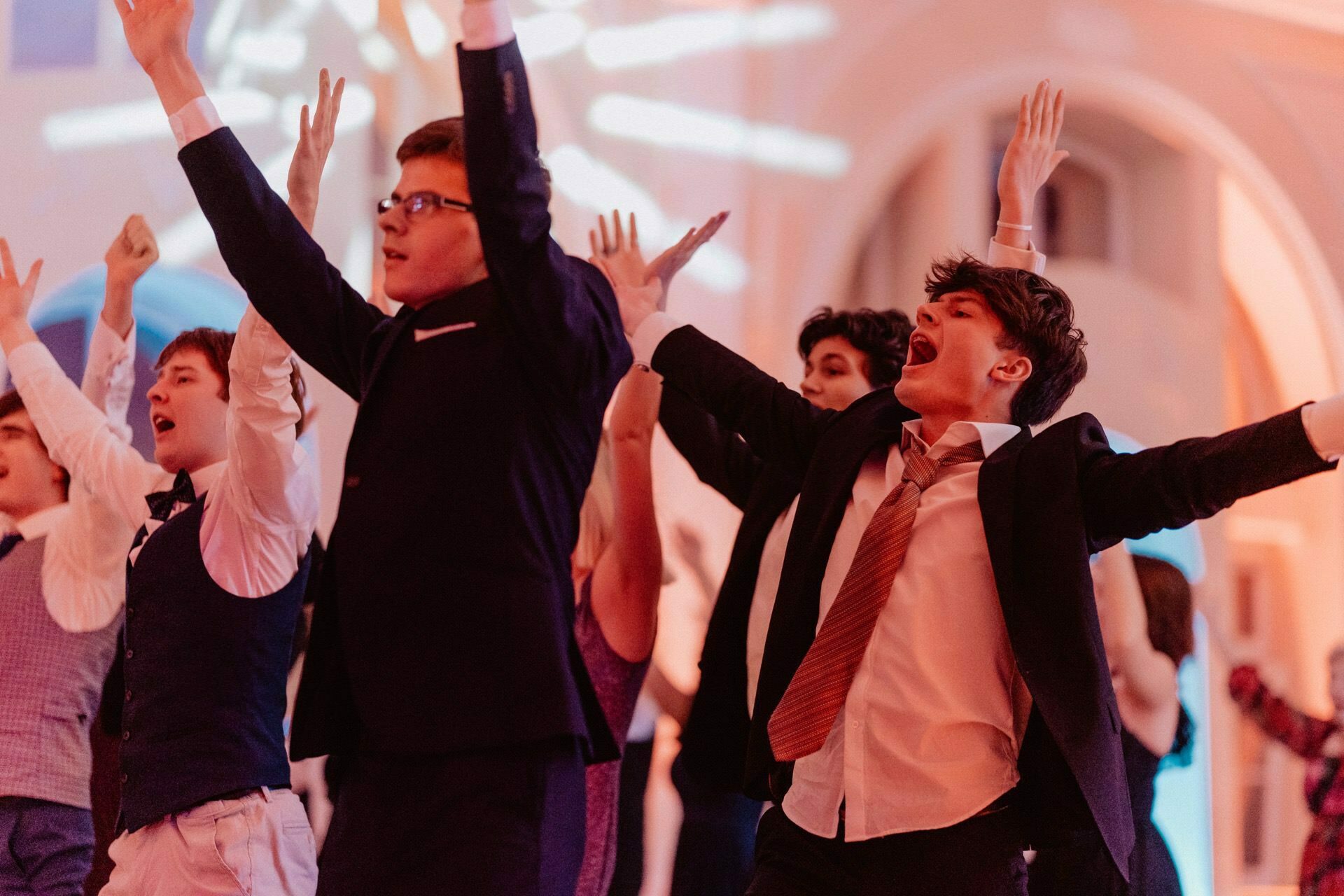 A group of formally dressed young adults enthusiastically dance with their hands raised, looking happy and energetic. There is warm lighting and an elegant interior in the background, suggesting a lively celebration or event, reminiscent of a vibrant prom reportage. 