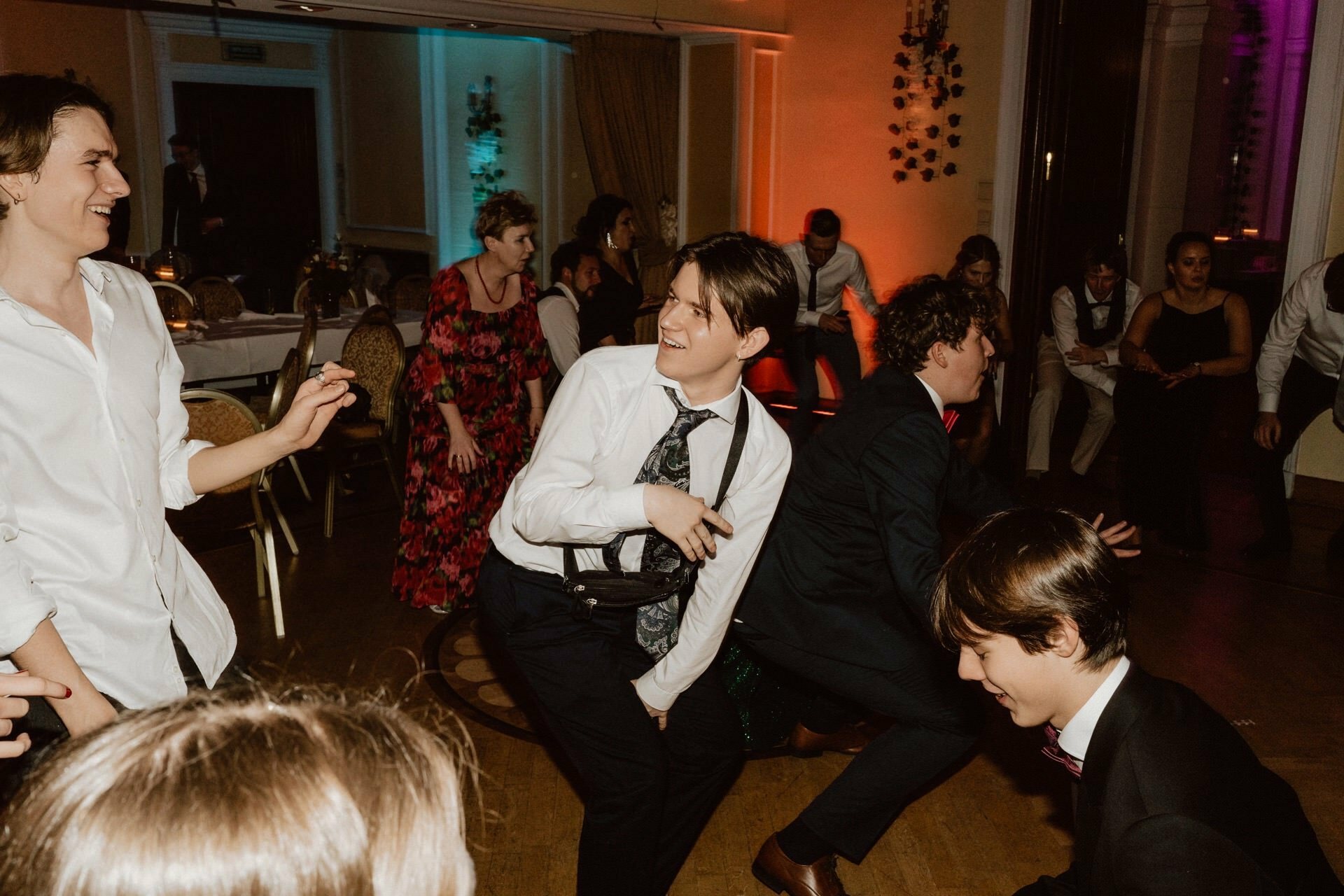 A lively celebration in a room with dancing people, reminiscent of *a prom reportage*. A few young men in white shirts and ties are in the foreground and dancing vigorously. A mix of people in formal attire, including suits and dresses, socialize, talk and dance. Colorful lights enhance the festive atmosphere.   