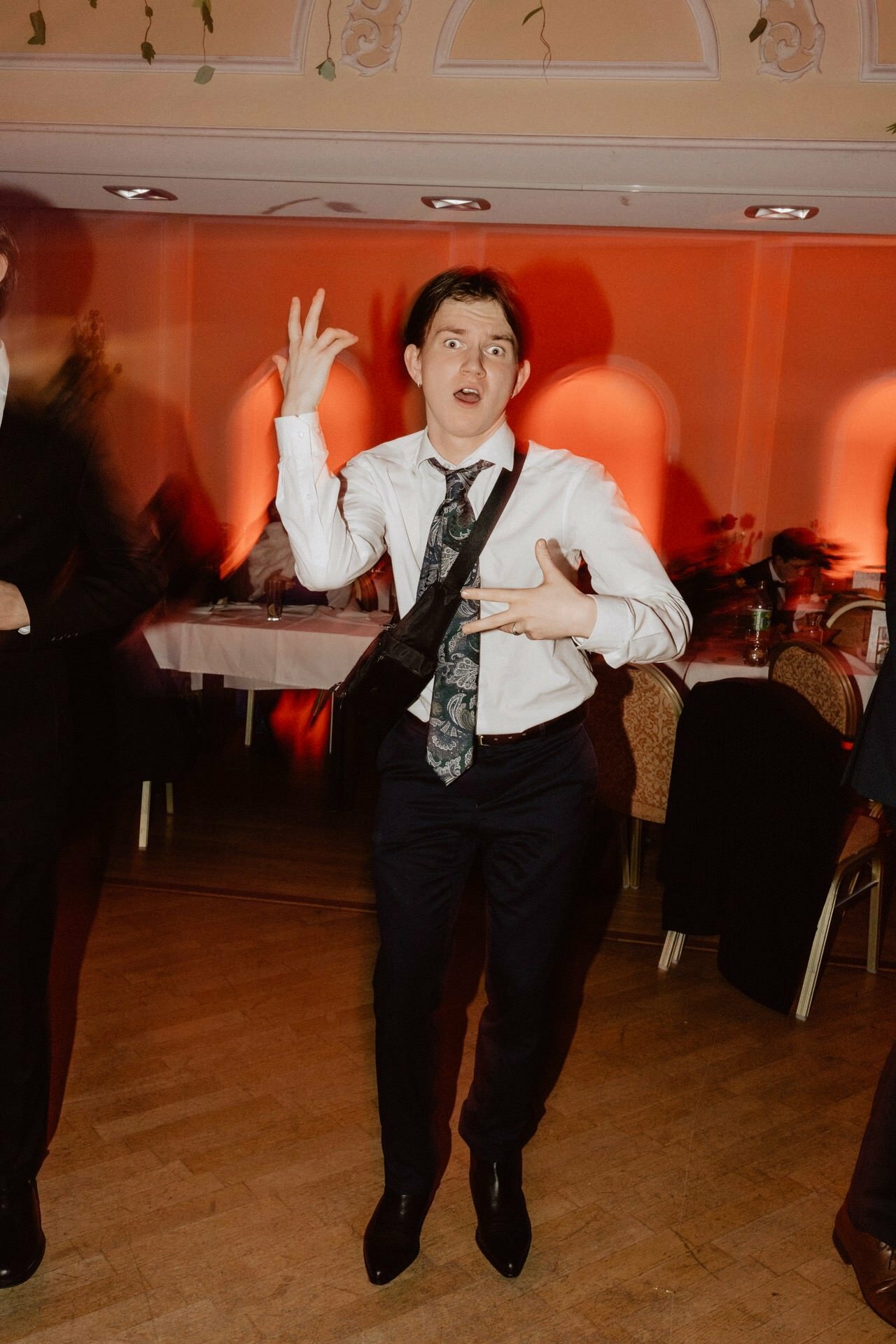 A young man in a loose tie and unbuttoned jacket moves expressively on the dance floor during a party in a room, reminiscent of a live reportage of a prom. The background is softly illuminated by the red-orange glow of wall sconces, and tables and chairs can be seen behind him. 