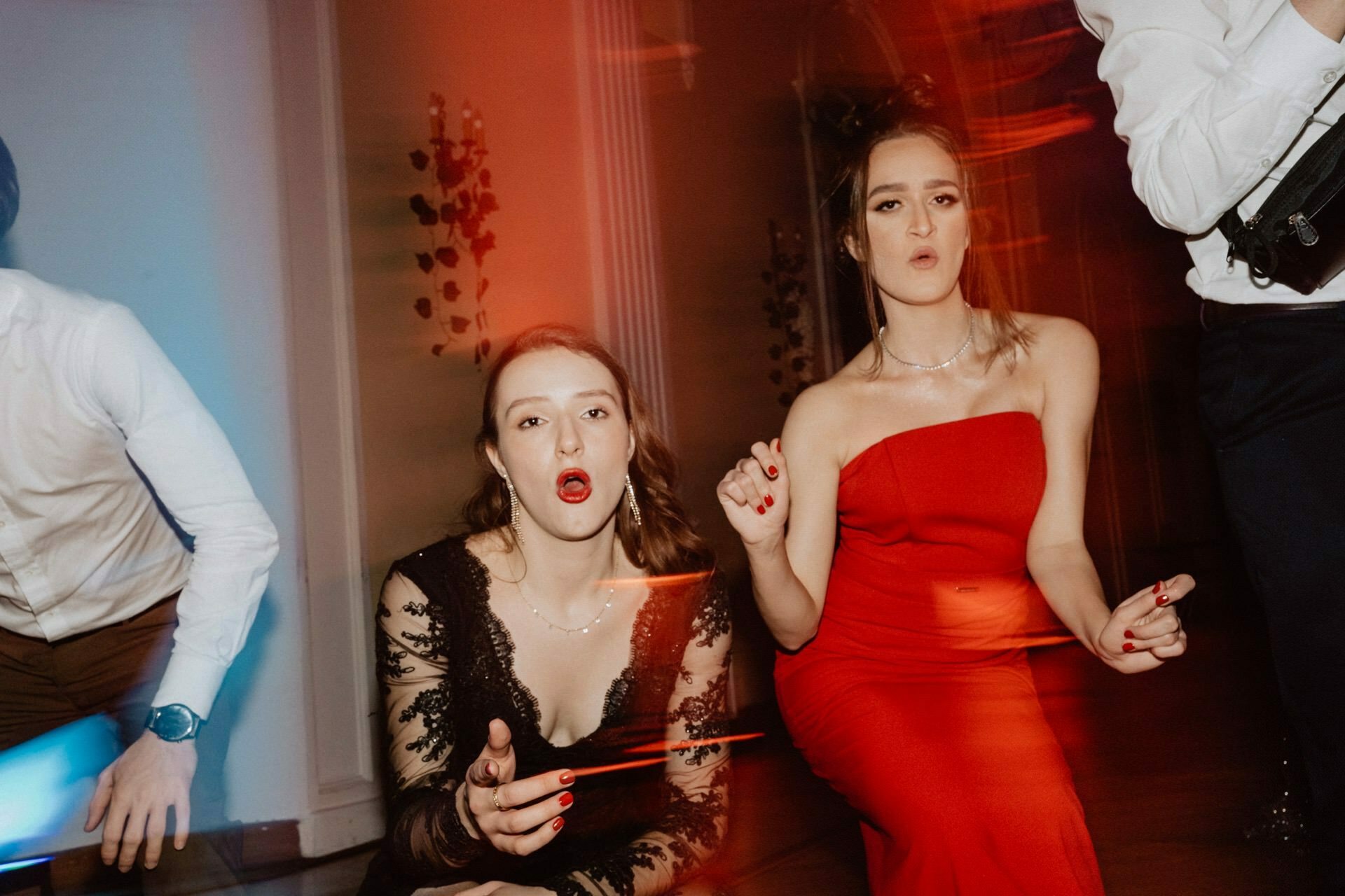 Two young women are sitting in a room, looking straight into the camera. One is wearing a black lace dress and the other a red dress. Both have expressions of surprise or excitement. The scene has red and blue lighting effects, capturing a vivid moment reminiscent of a prom reportage.   