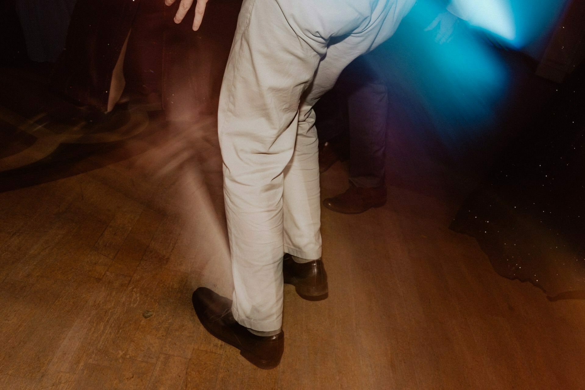 A person wearing light-colored pants and dark shoes dances on a wooden floor, which resembles a vivid reportage from a prom. The motion blur captures the dynamic movement as blue and orange lights create a lively atmosphere around the dancer's legs. 