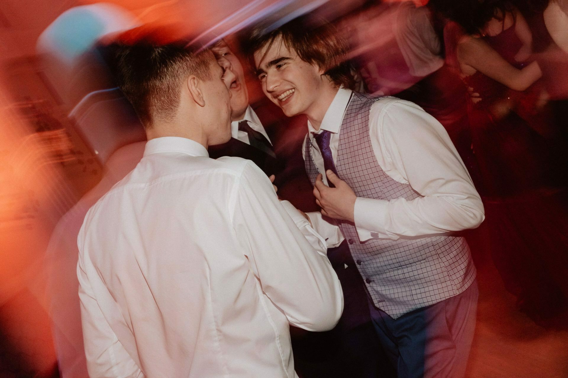 Three young men chatted during a lively dance party, all dressed in formal attire. The image captures a blur of motion, suggesting movement and possibly dancing. The background is filled with colorful lighting and other participants, giving the atmosphere of a bustling prom reportage.  