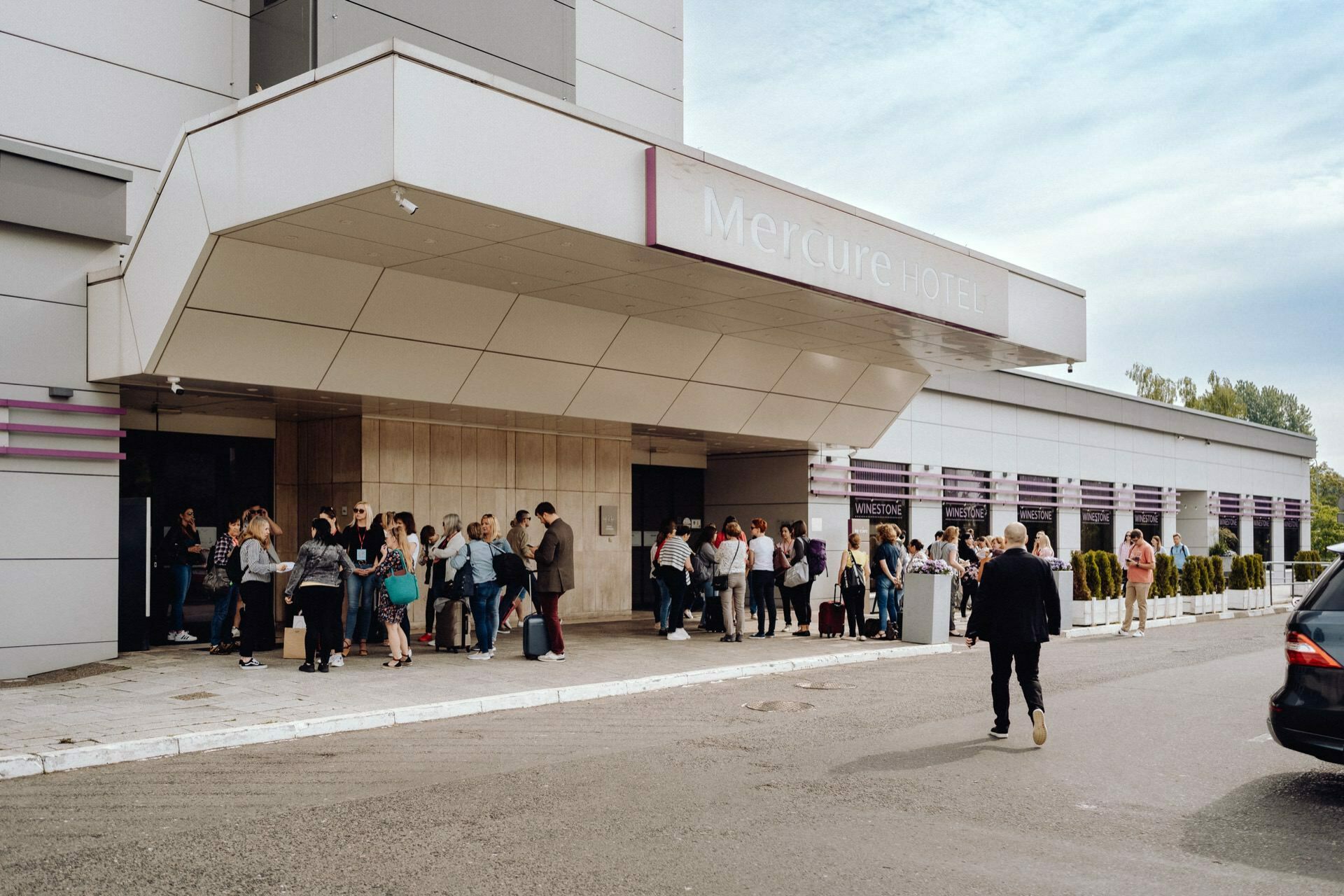 In front of the entrance to the Mercure Hotel stands a large group of people with luggage, talking and waiting. One person walks away from the hotel entrance. The modern building with its gray and white facade contrasts beautifully with the blue sky, perfect for an event photographer Warsaw to capture.  