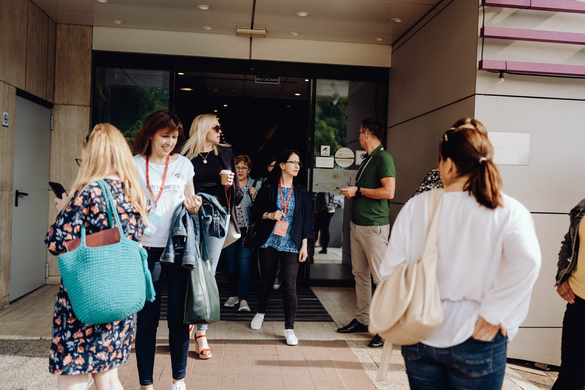 A diverse group of people exits a modern building through a glass door. Some are carrying bags, others are holding drinks, and some are engrossed in conversation. The scene, captured in daylight by event photographer Warsaw, is characterized by a casual, lively atmosphere.  