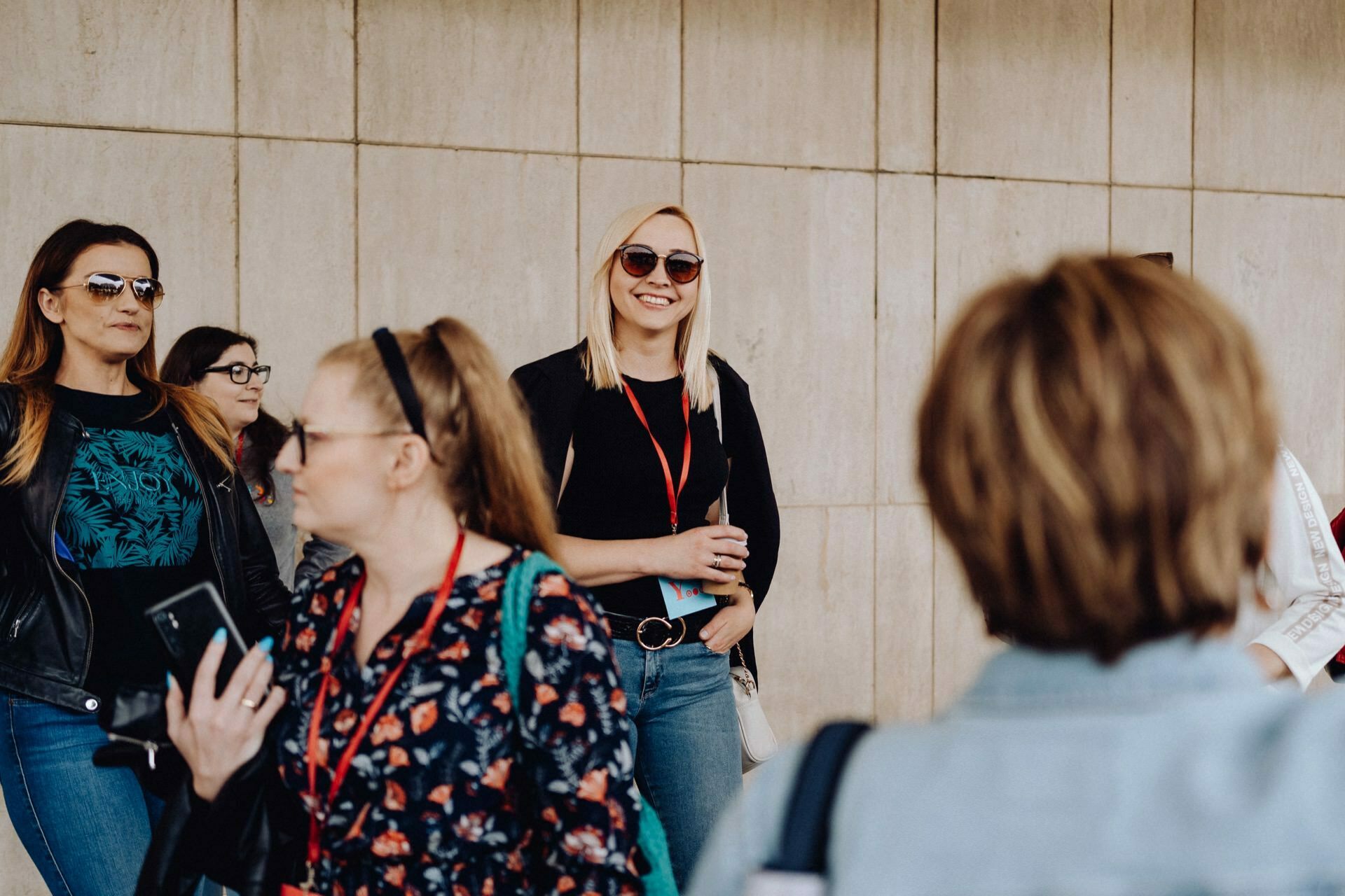 A group of people, mostly women, are standing and walking together outside near a beige tiled wall. Most are wearing casual clothes and sunglasses. The blonde in the middle smiles directly at the camera while holding a drink. Around their necks hang red lanyards - a vivid photo essay of the event, full of vibrant energy.   