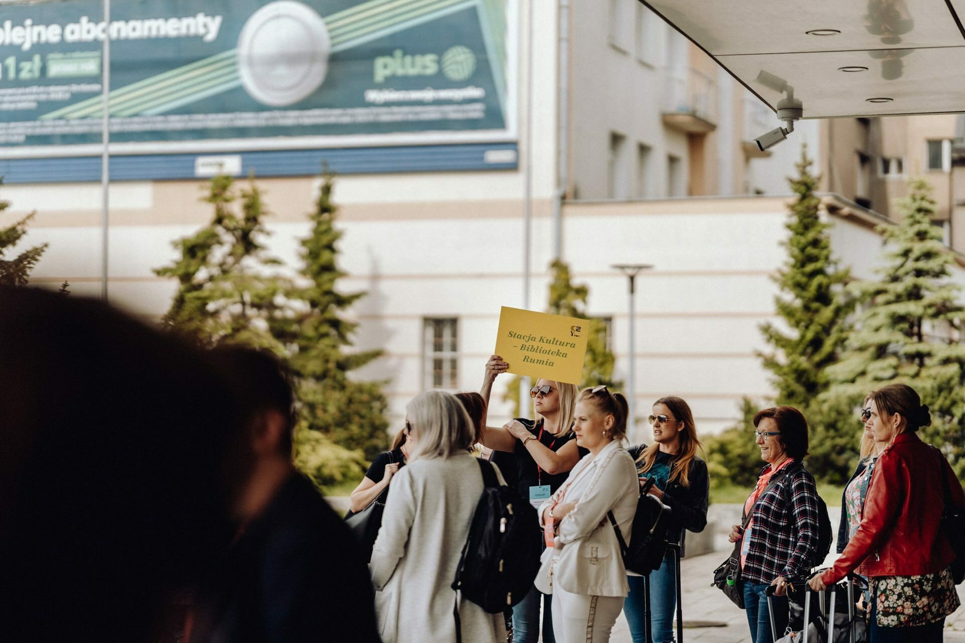 A group of people gather outside. One person prominently holds a yellow sign that reads "Ešte Stojím a bojujem." The group stands under a canopy, while trees and buildings can be seen in the background. This scene can easily be captured by any skilled photographer at the event.   