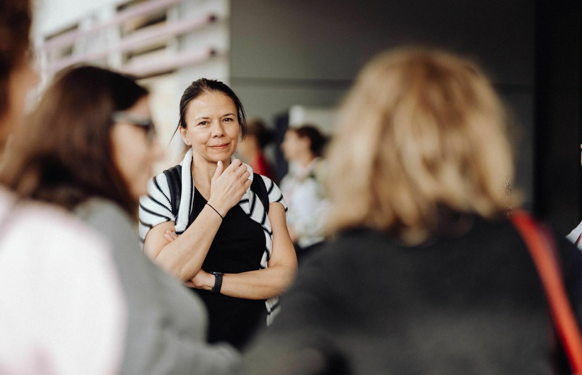 In the group stands a woman with long dark hair, wearing a black shirt and striped cardigan. She smiles and rests her hand under her chin, looking at someone outside the frame. This photo report from the event shows a slightly blurred background with several people talking.  