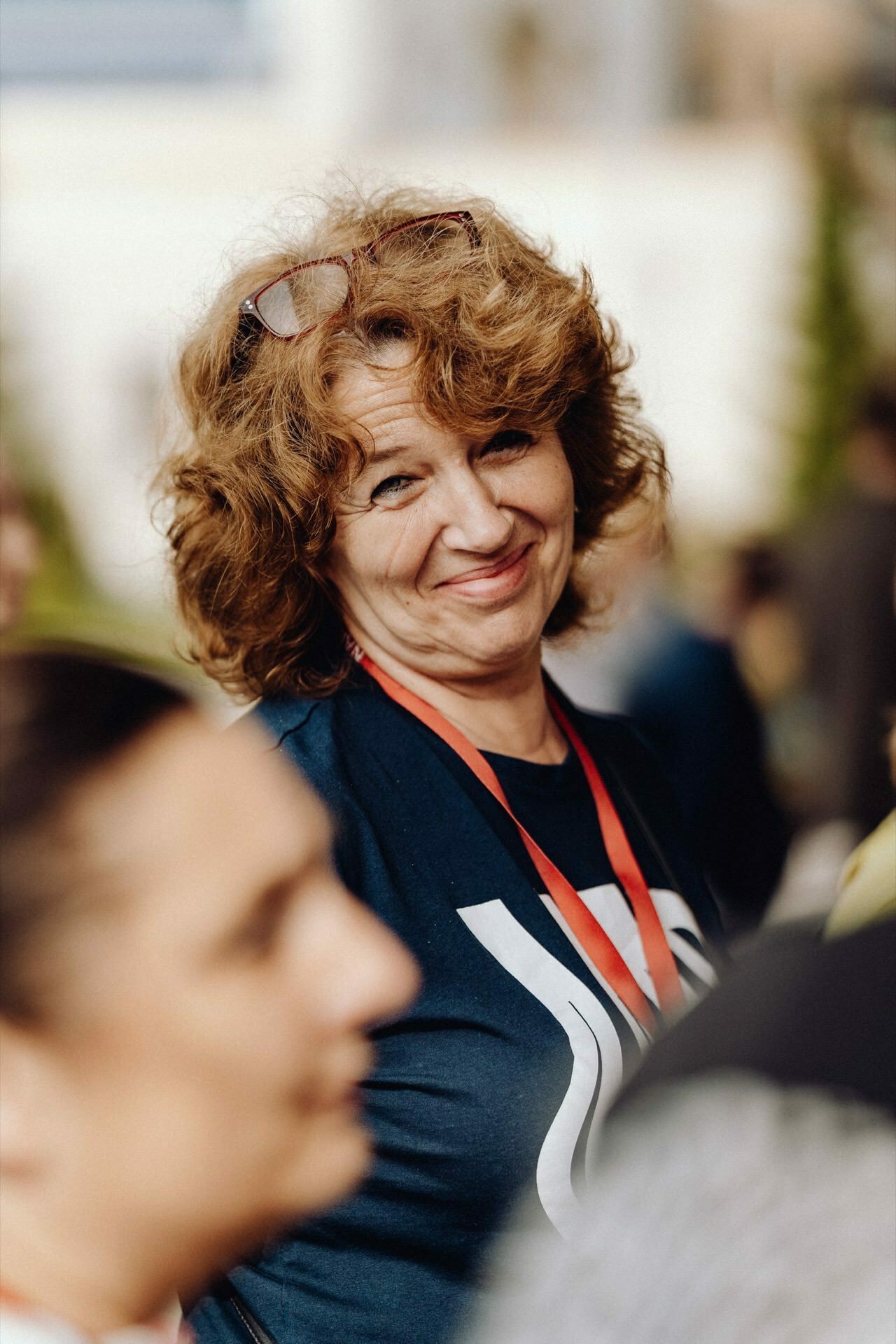 A woman with curly hair and glasses on her head smiles at the camera. She is wearing a dark shirt and a red lanyard. The background is blurred, capturing the essence of the photojournalism of the event, while the other person in the foreground remains out of focus.  