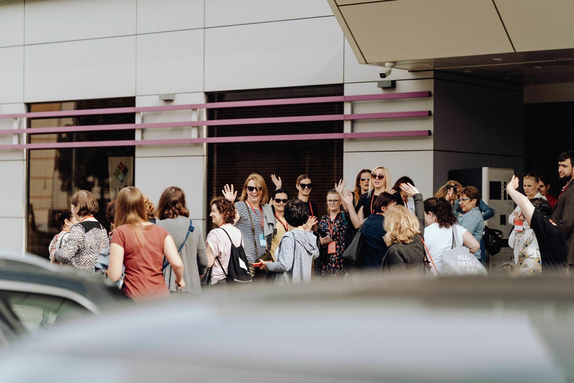 A group of people gathered outside the building, and some waved toward the camera. The building has a modern facade with white panels and purple accents. Various cars are partially visible in the foreground. The atmosphere captured by event photographer Warsaw seems lively and vibrant.   