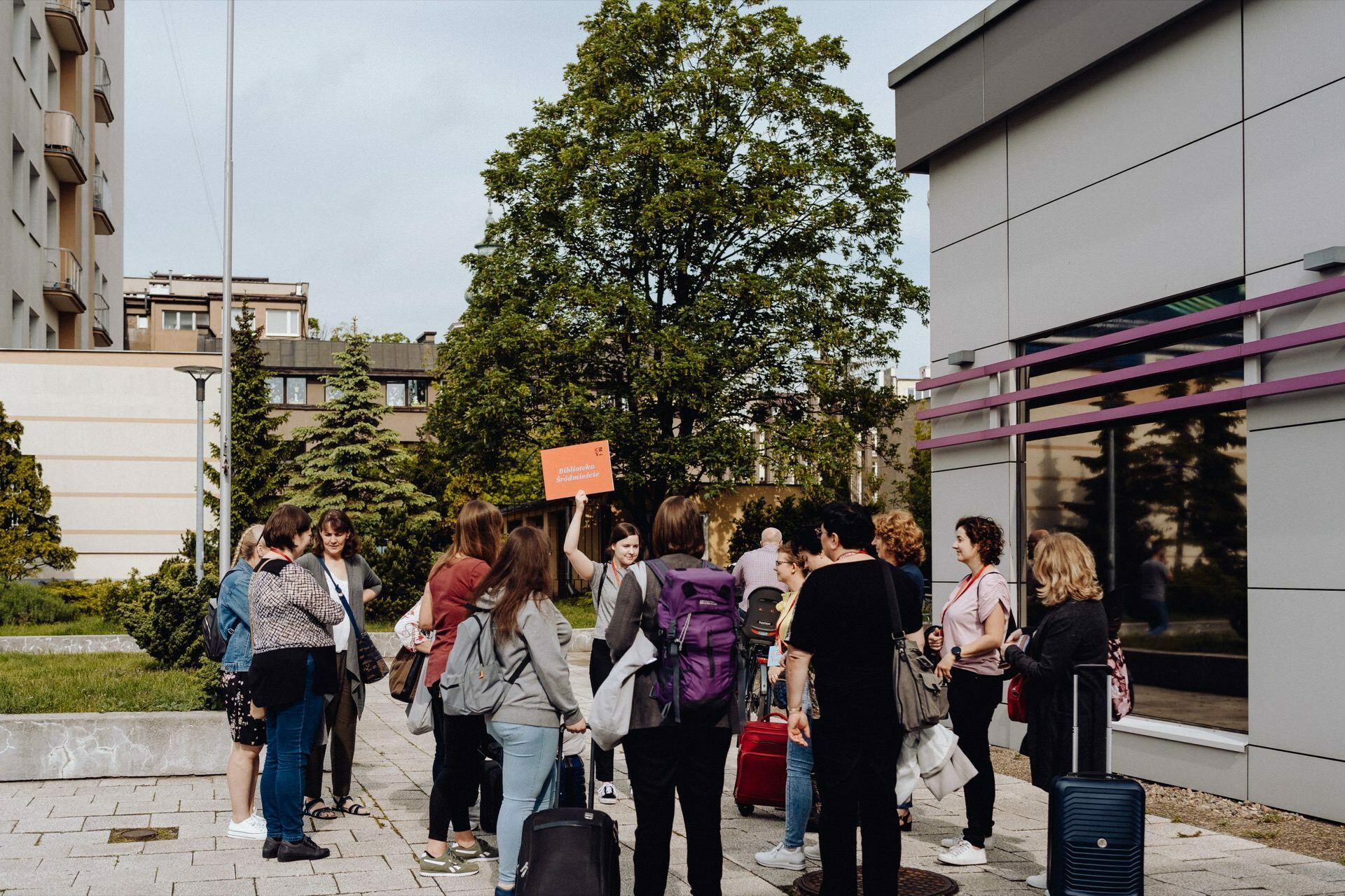 A group of people are standing outside near the building, and one person is holding an orange sign and probably participating in a tour or gathering. Luggage is visible and trees are in the background. The urban scene suggests that this may be part of a photo essay of an event by a photographer from Warsaw.  