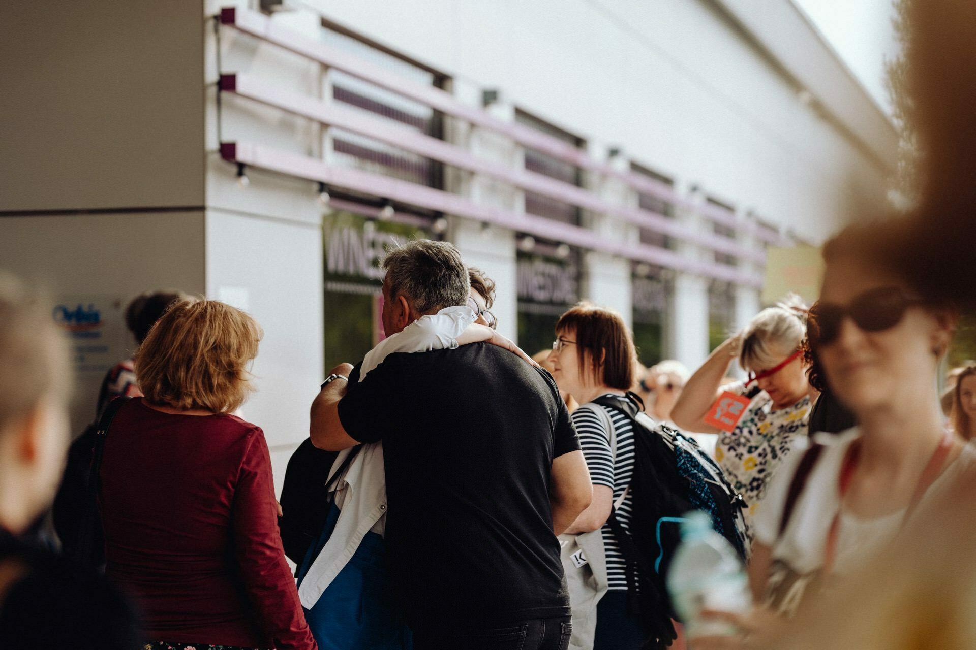 A crowd of people in front of the building, some immersed in conversation. In the foreground, a man in a black shirt hugs another person dressed in white. In the background you can see a modern building with windows and purple accents, beautifully captured by a photographer from Warsaw as people walk past.  