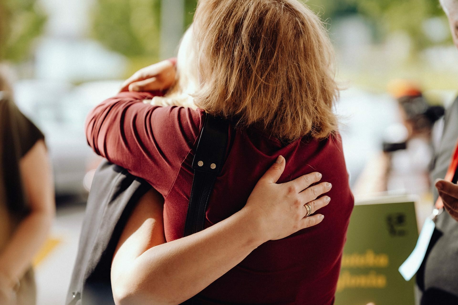 Two people embrace warmly in an outdoor setting, beautifully captured by a photographer from Warsaw. Only the upper body and arms are visible, with one person's hand gently resting on the other's back. The background is blurred, alluding to the greenery and blurred figures.  