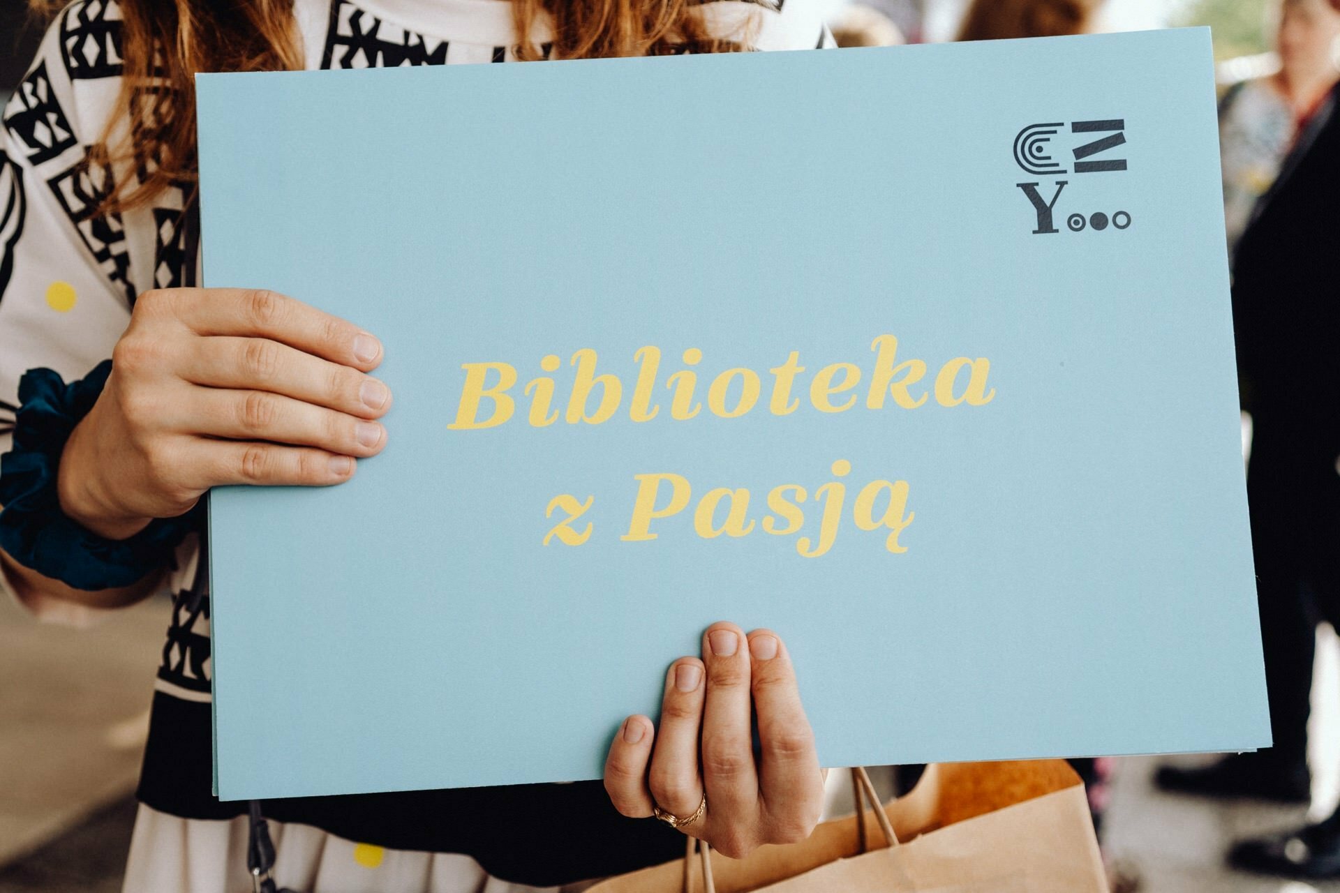 An individual holding a bright blue placard with the yellow text "Library with Passion," which in English means "Library with Passion." Captured by event photographer Warsaw, the individual is wearing a patterned outfit with black and white designs. 