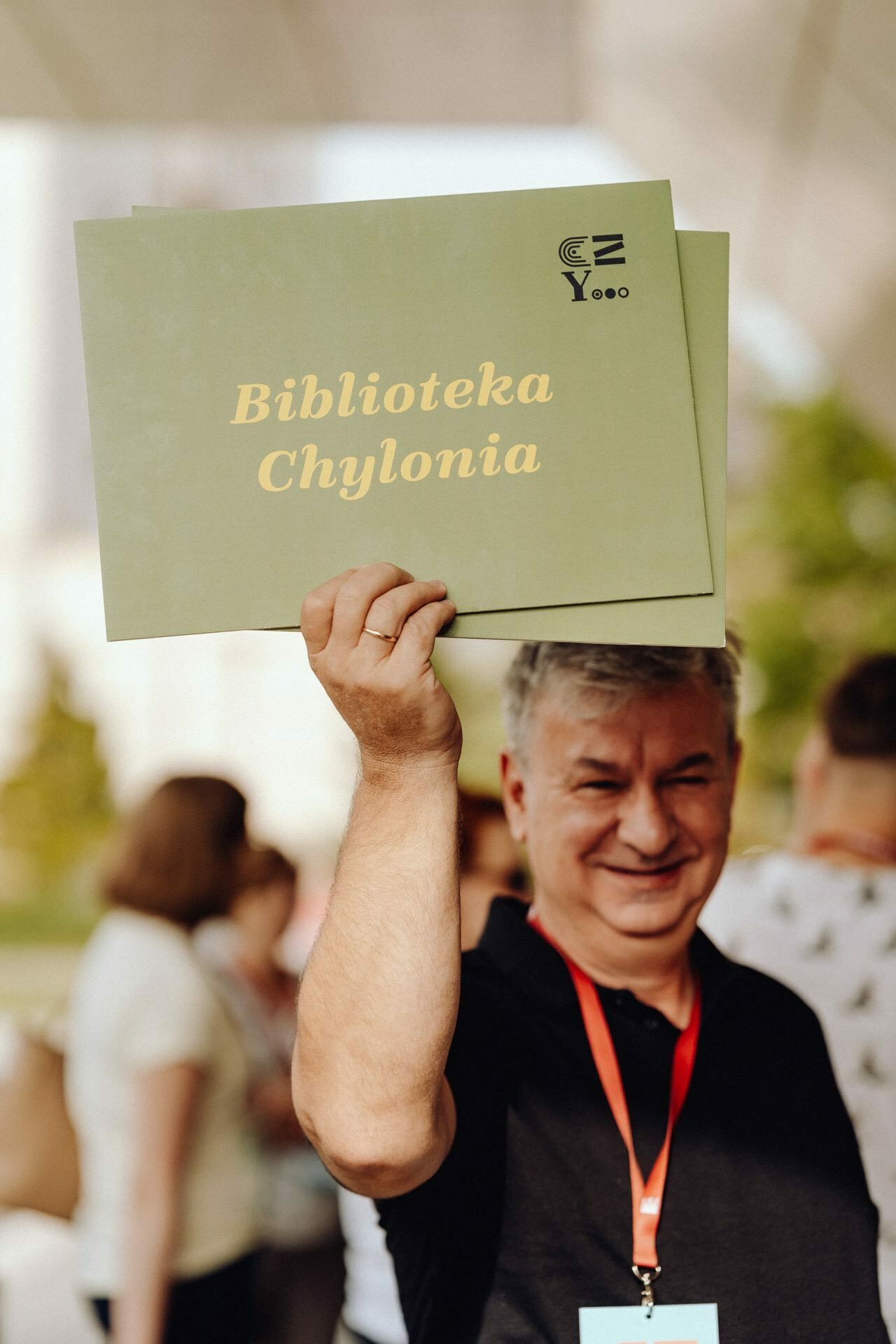 The smiling person captured in the *photograph from Warsaw* is holding up a green sign with the words "Chylonia Library" in yellow text. They are wearing a black shirt and a red lanyard. In the background you can see blurred people and greenery.  