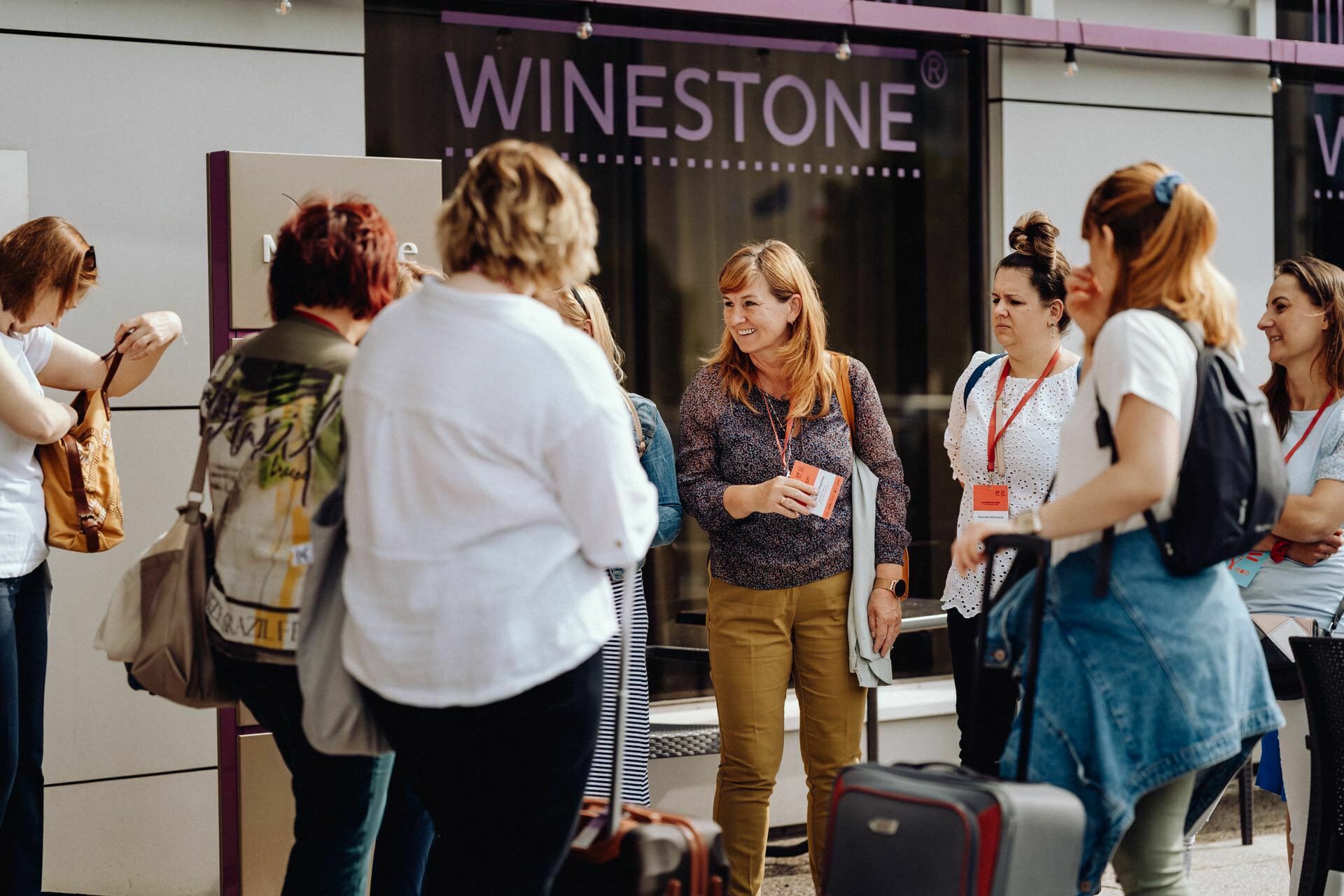 A group of people stands in front of a building with a sign reading "WINESTONE." A woman in the middle, holding a brochure, addresses the group. Some have lanyards suggesting a conference or tour. Others are carrying luggage, and all appear to be immersed in conversation - perfect for any *Warsaw photographer* to capture the moment.   