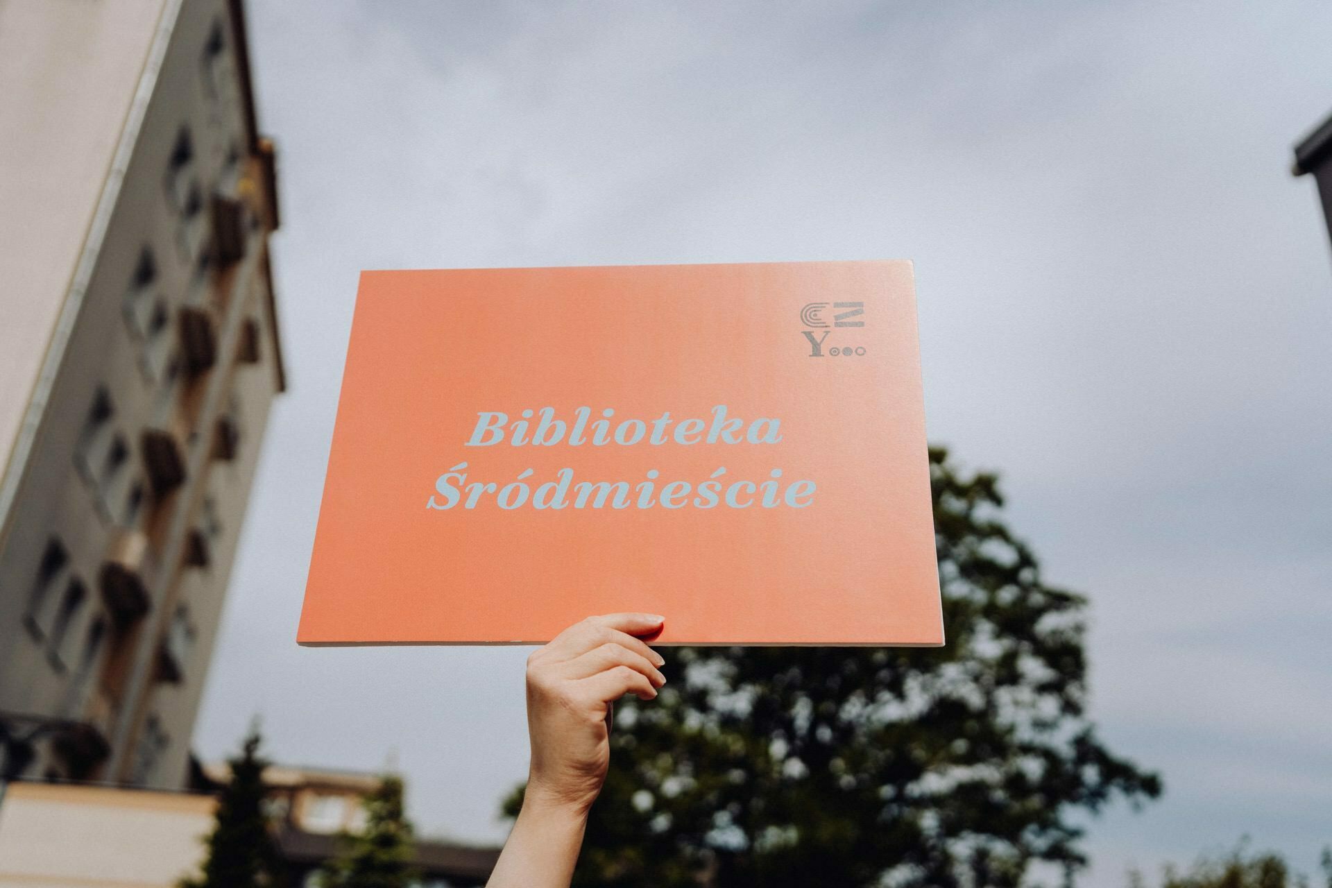 The hand holds a coral-colored plaque with the white text "Downtown Library" against a background of a tall building, green trees and cloudy skies - a perfect photo by a photographer from Warsaw.