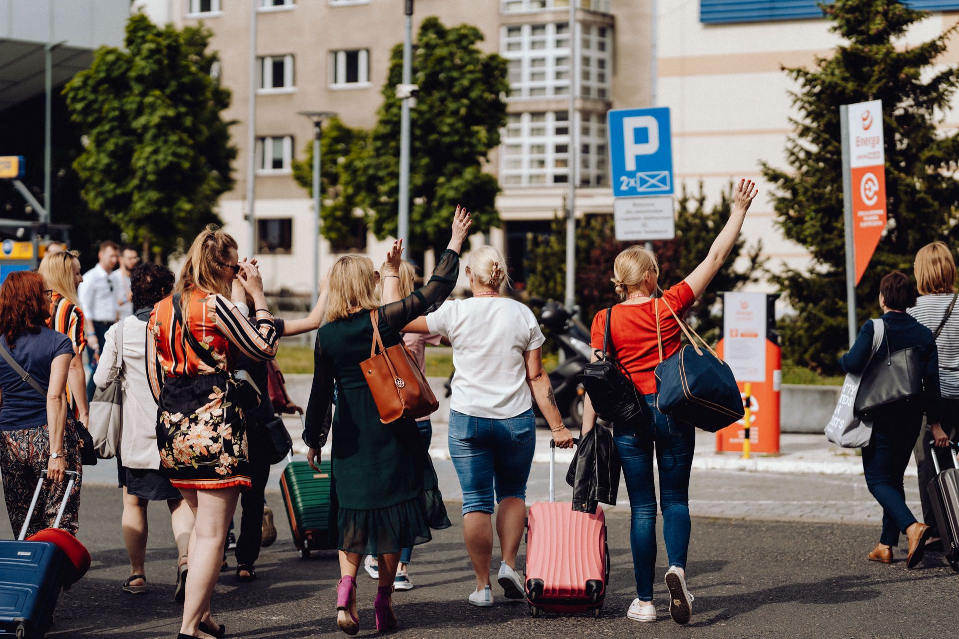 A group of people with luggage walk towards the building, waving their hands. The parking sign can be seen on the street next to various buildings and trees in the sunny background. This moment can be beautifully captured by a photographer for an event as part of a great photo essay of an event in Warsaw.  