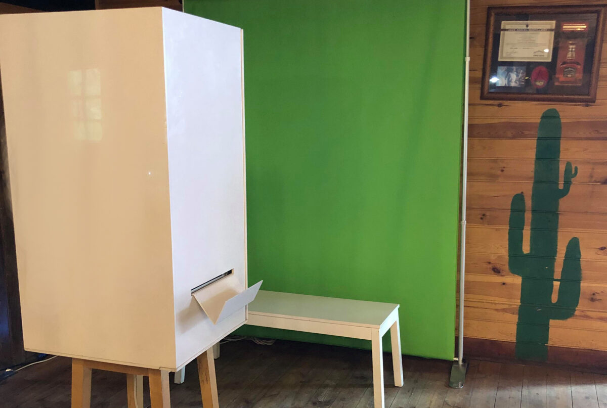 A white photo booth with a small opening at the bottom stands in front of a green screen, next to a white bench. On the wooden wall to the right hangs a framed testimonial by Marcin Krokowski, an event photographer in Warsaw. A painted green cactus is visible on the wooden wall.  