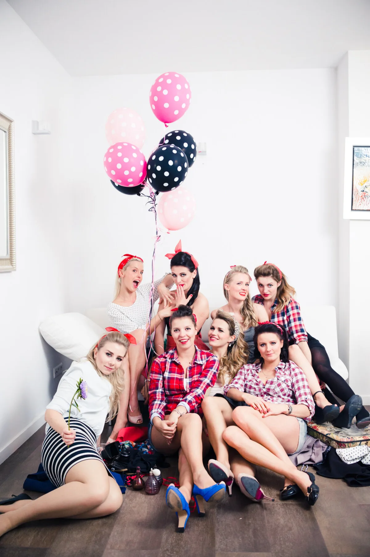 A group of nine women dressed in retro 1950s-style outfits with bright red lipstick pose in a room. Marcin Krokowski captured the scene perfectly, as most of them wear plaid shirts, shorts or high-waisted skirts and sport bandanas. One woman in the back holds balloons with pink and black dots.   