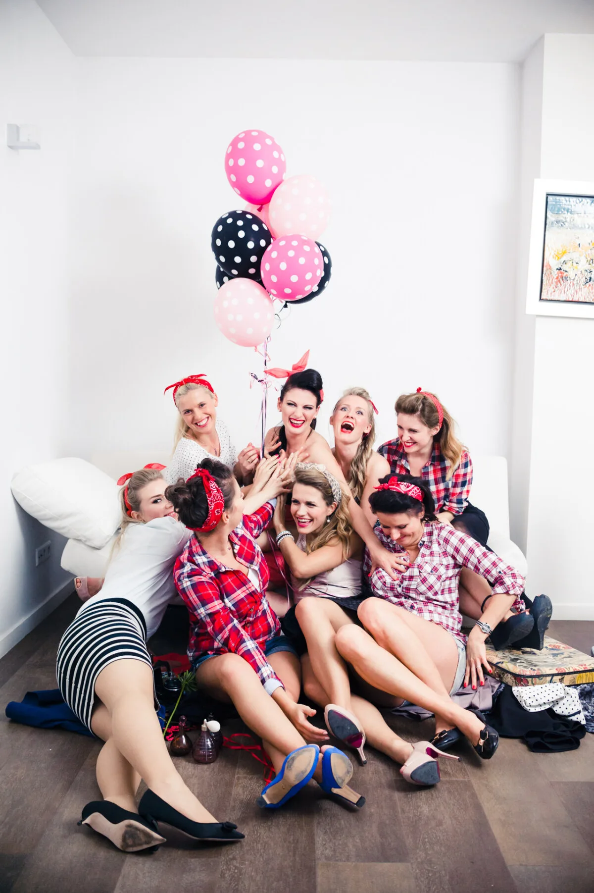 A group of eight women dressed in retro-inspired clothing sit joyfully on the floor, clustered around two women holding pink and black polka dot balloons. They appear to be celebrating in a bright white room, with a photo hanging on the wall in the background, expertly captured by event photographer Marcin Krokowski. 