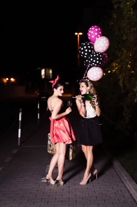 Two women in stylish outfits stand outside at night. The woman on the left, in a pink dress, holds a glass of wine. The woman on the right, in a black skirt, holds colorful balloons and drinks from a bottle. They both seem to be celebrating and having a good time, which was perfectly captured by photographer Marcin Krokowski in Warsaw.   