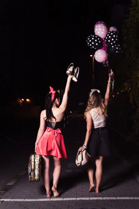 Two women seen from behind are walking down a dimly lit street at night. One holds high-heeled shoes and a suitcase, wearing a bright dress with a large bow. The other is holding polka dot balloons and a purse, wearing a black skirt and a bright top - perfectly captured by Marcin Krokowski, a photographer in Warsaw.  