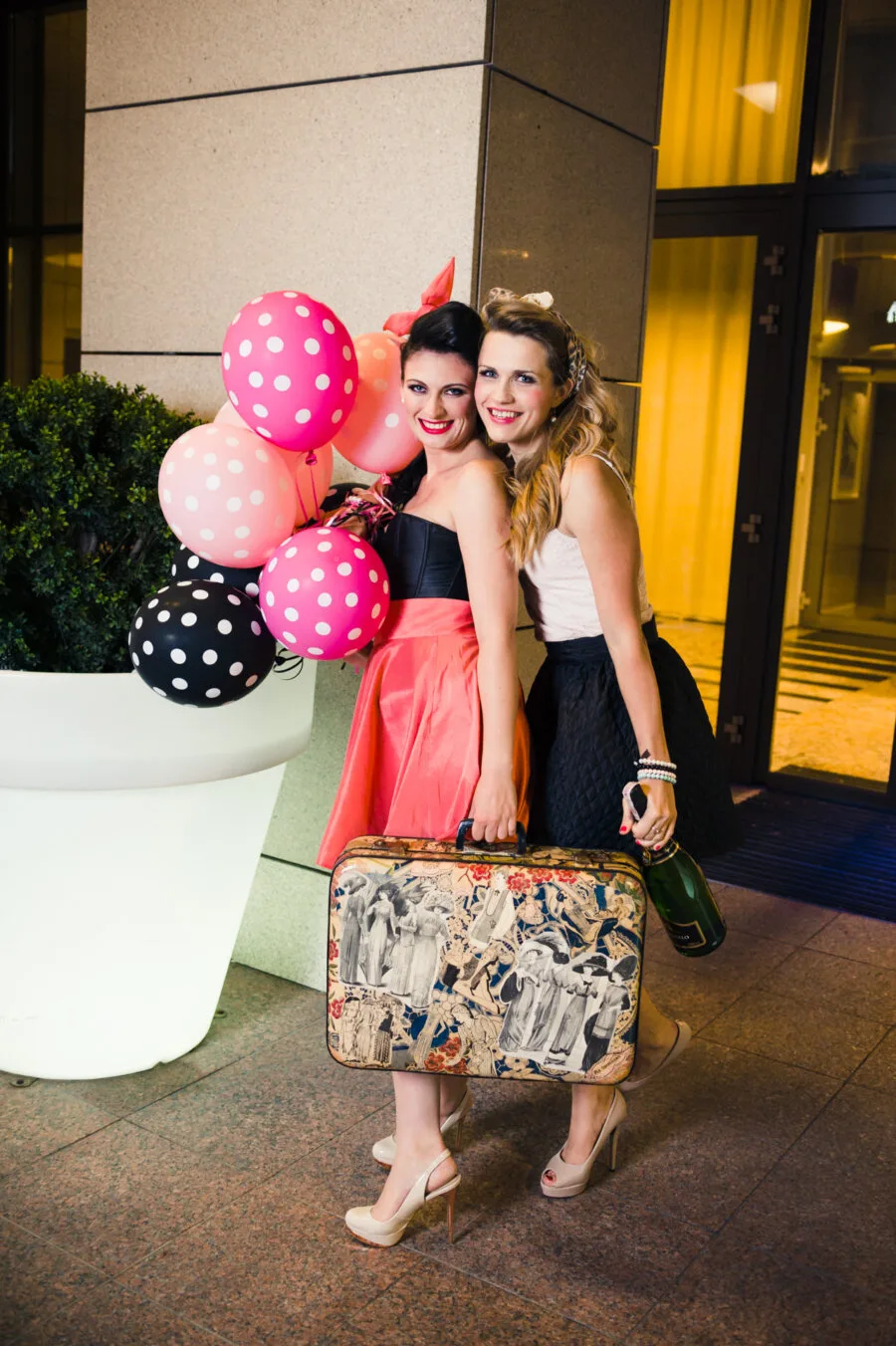 Two smiling and stylishly dressed women are standing close together. One is wearing a red dress and holding colorful polka dot balloons. The other is wearing a black and white dress and holding a decorative suitcase and a bottle. They are standing outdoors, next to a large white pot, in a night scene photographed by Marcin Krokowski.   